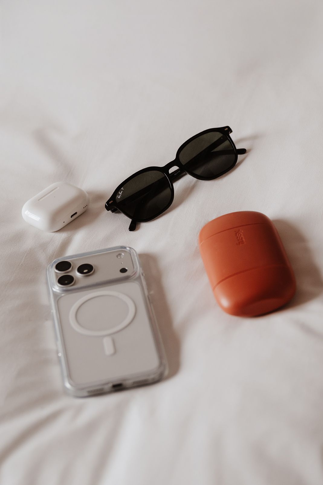 Flatlay of iPhone with MagSafe case, sunglasses, AirPods case, and brown leather case on white bedding.
