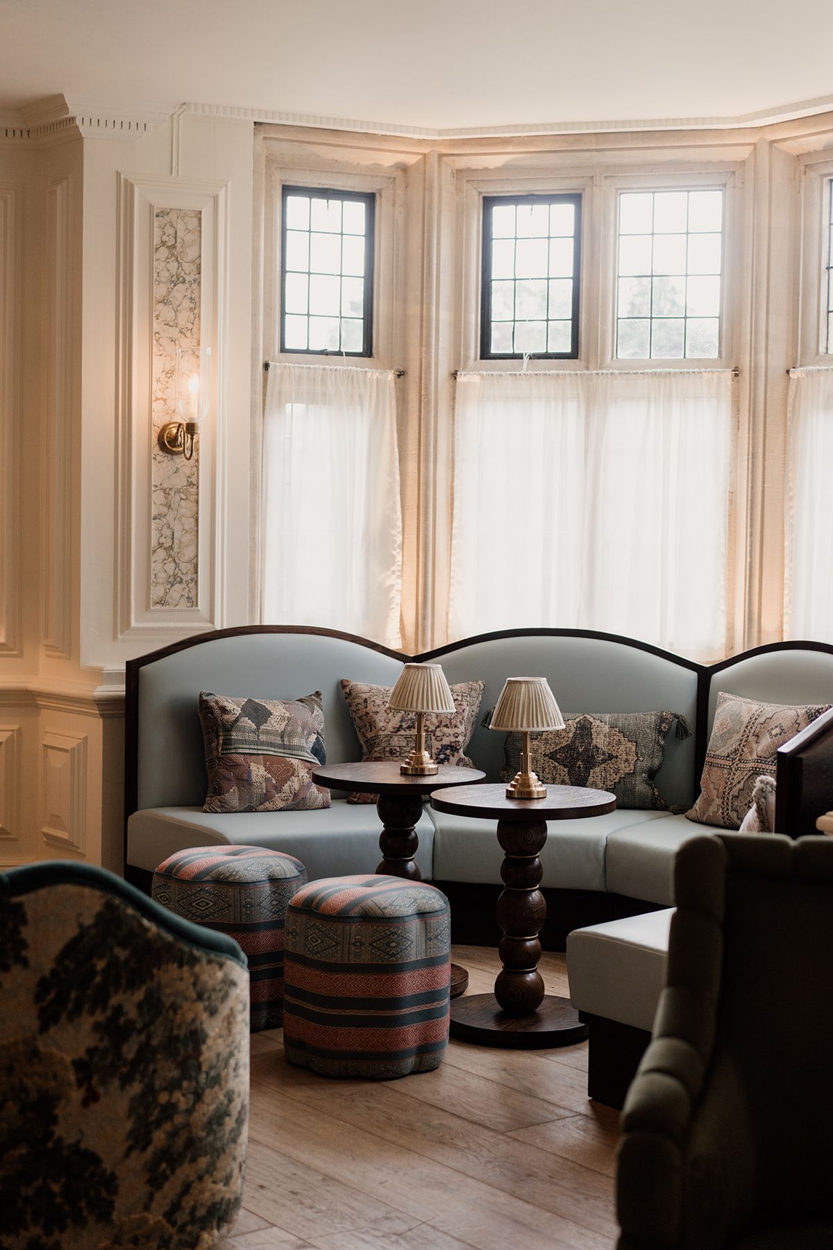 Cozy traditional lounge with teal curved sofas, plaid ottomans, table lamps, and leaded bay windows.