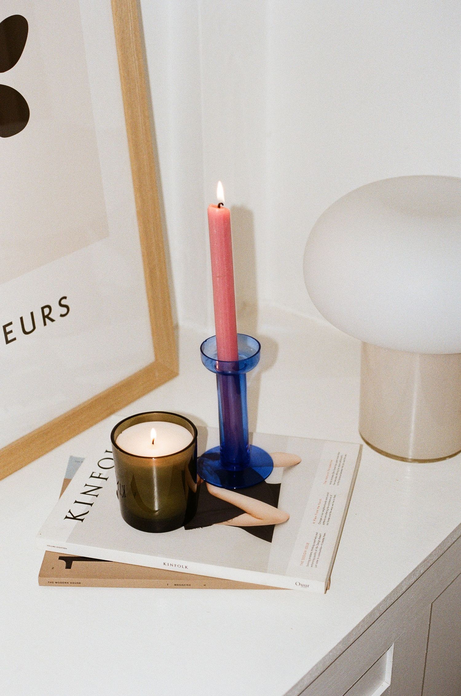 Minimalist home decor with lit pink taper candle in blue holder, dark green scented candle, and lamp on white surface.