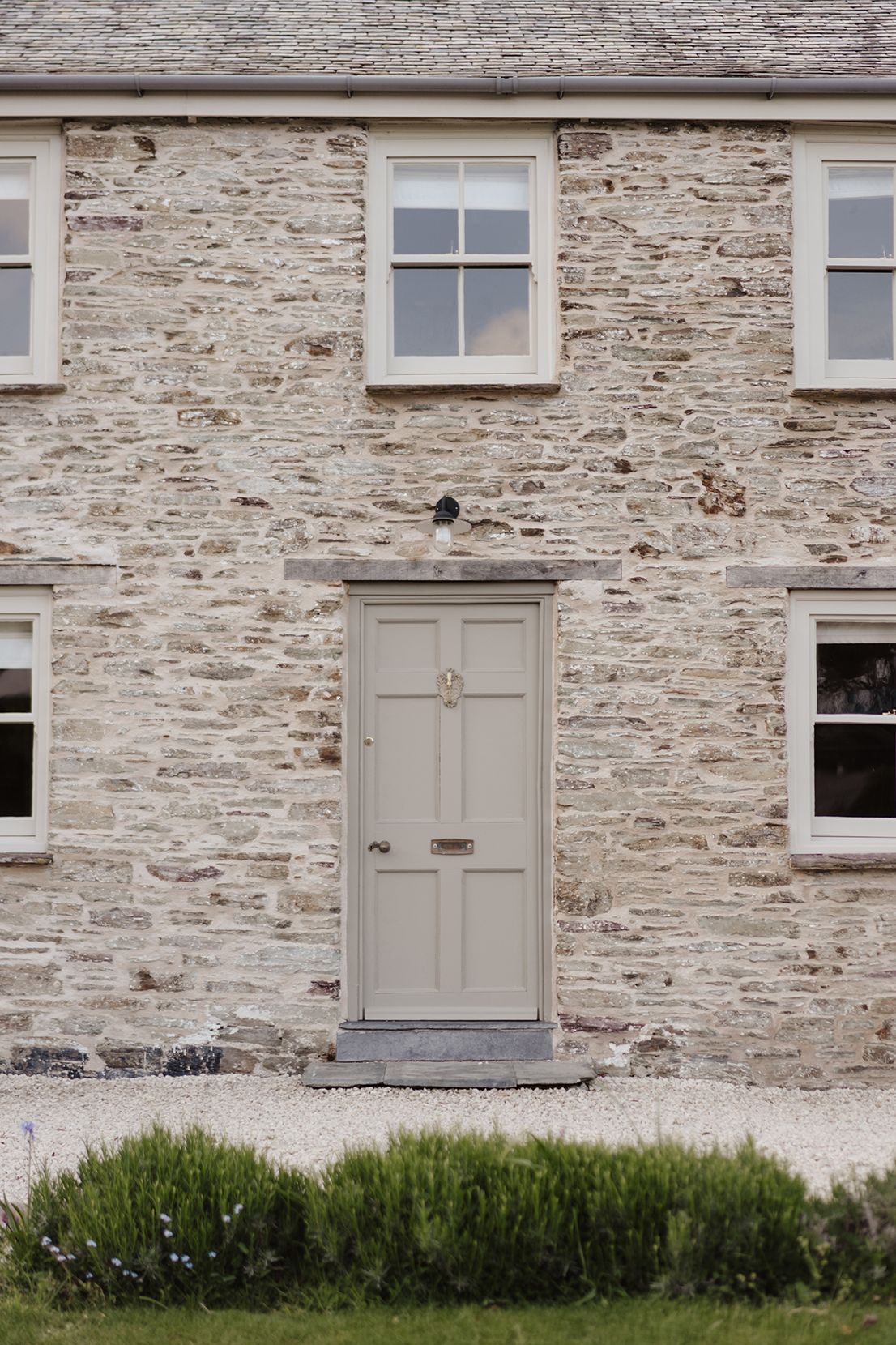 Traditional stone cottage facade with taupe painted door, white sash windows, and rosemary border planting.