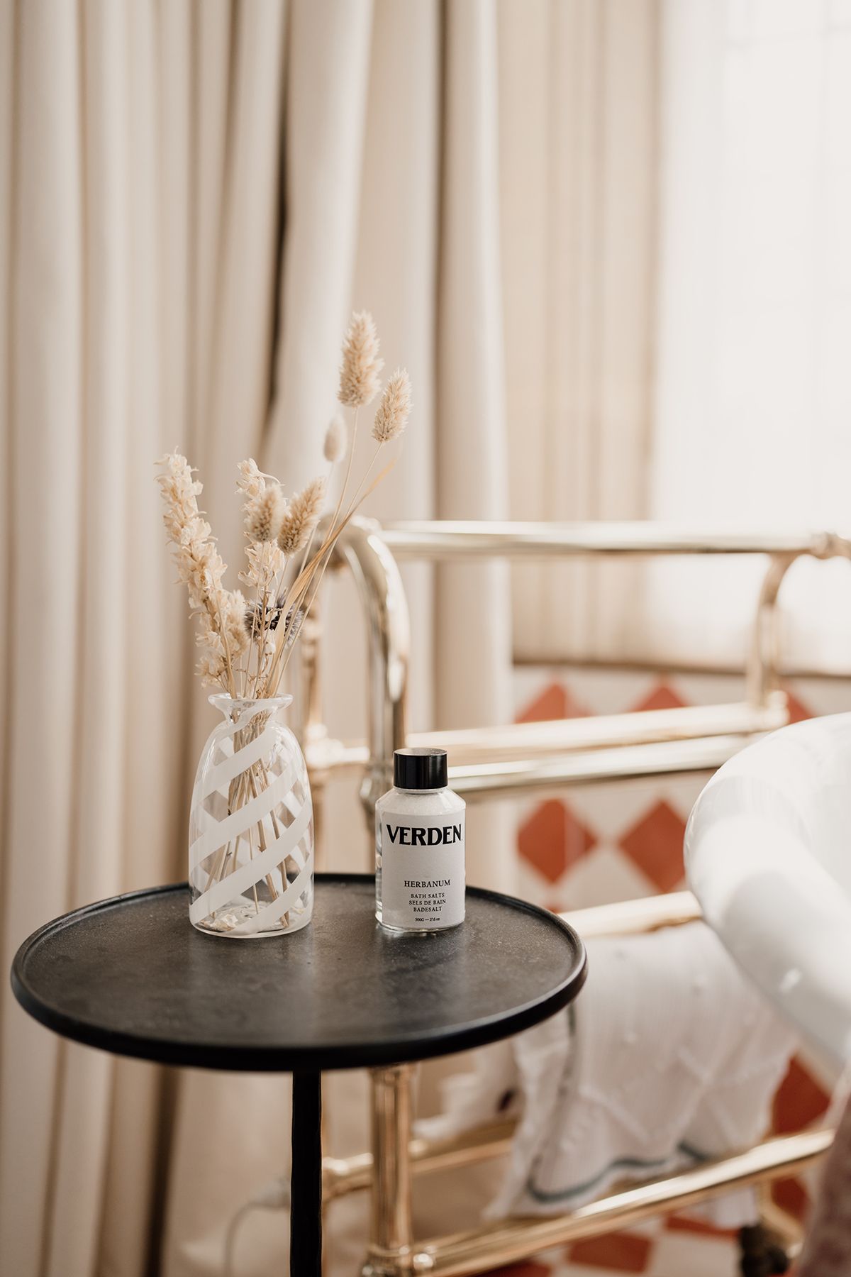 Verden aromatherapy bottle on dark side table beside dried grass vase near a gold bed frame with curtains.