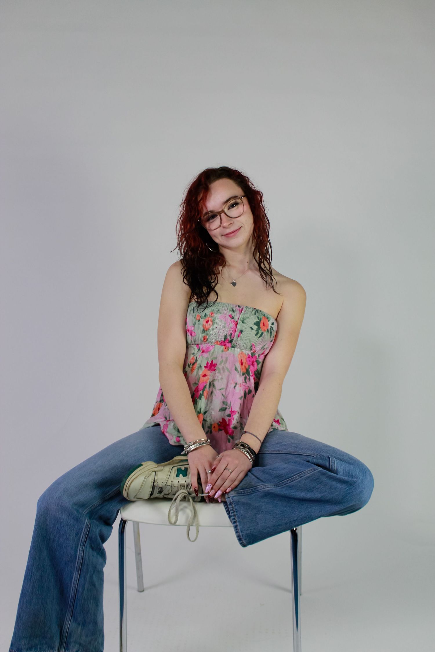 Studio portrait of a woman posed sat causally on a chair. Fashion photo. Wearing her red hair down, jeans and pink floral top.