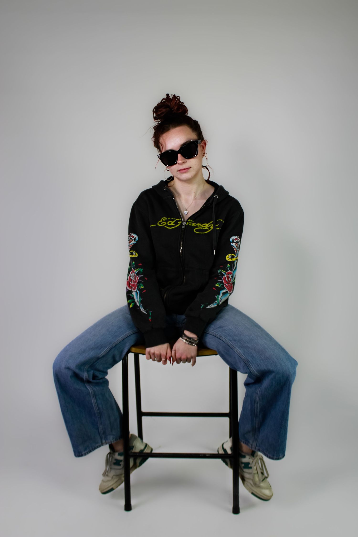 Studio portrait of a posed sat casually on a chair. Fashion photo. Wearing her red hair in a bun, jeans and a hoodie.