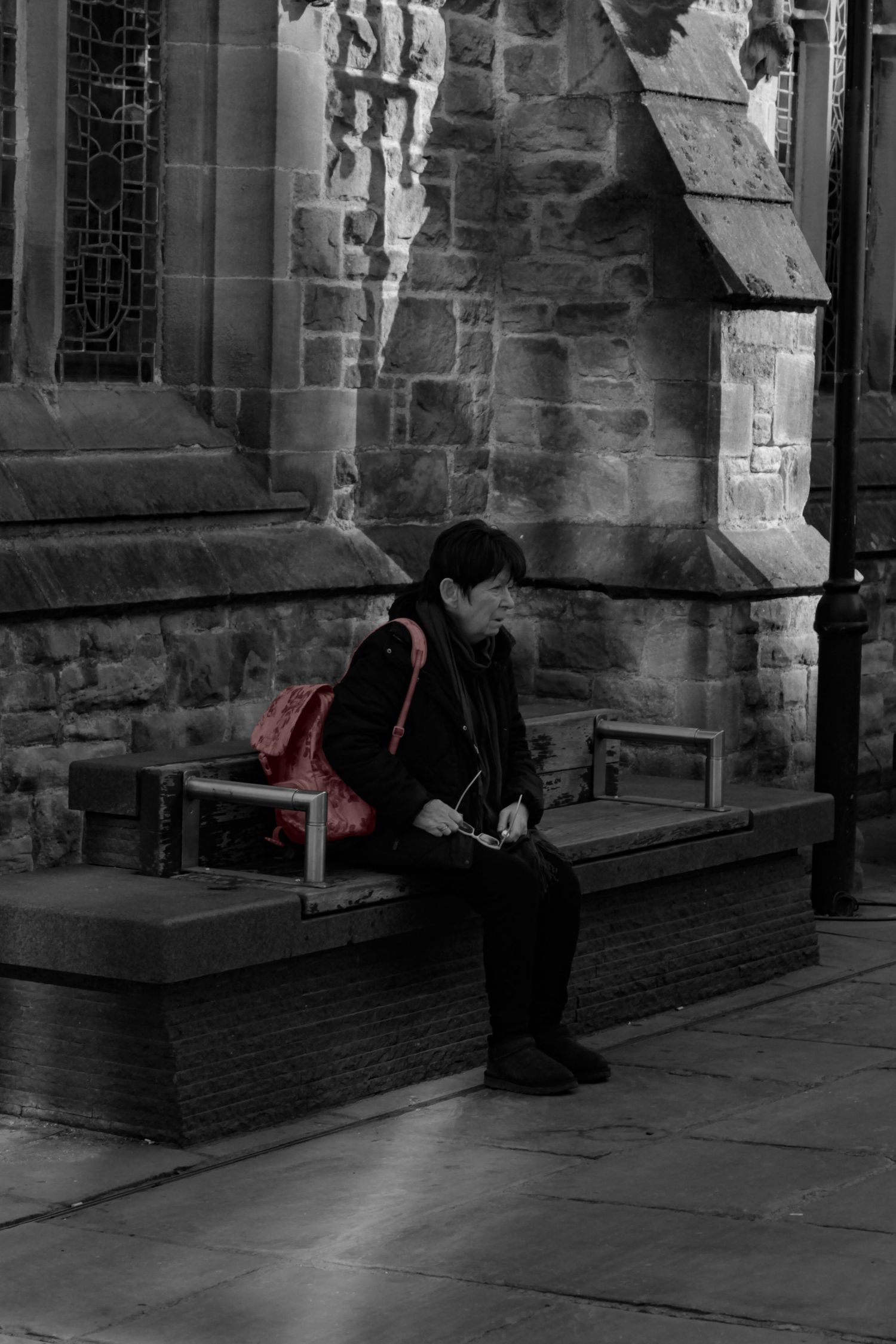Street photography. Mostly black and white photo of a woman sat on a bench. Her bag is red.