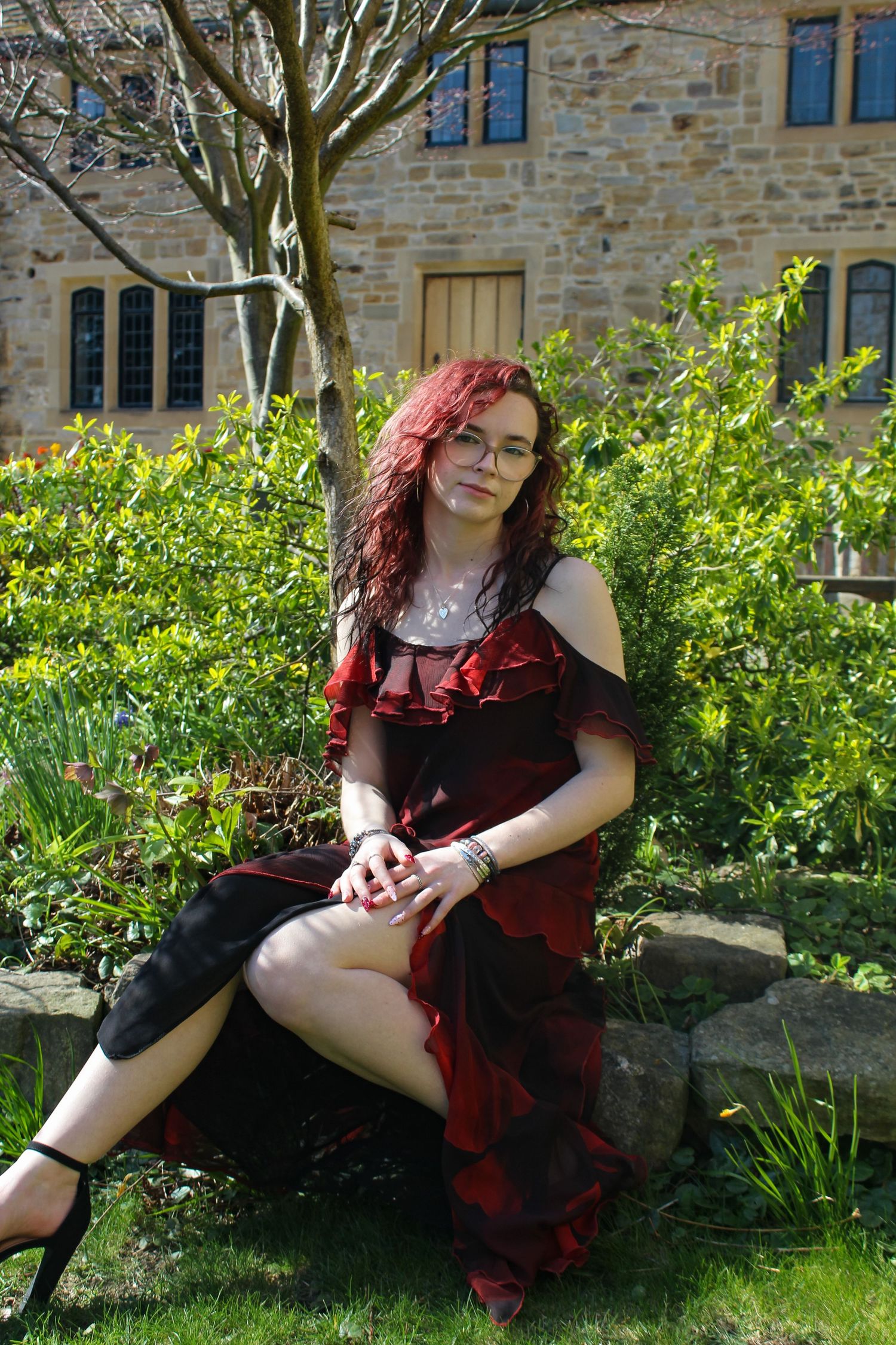 Editorial fashion portrait of a woman posed a garden with a beautiful background. Wearing her red hair down and a red dress with high heels.