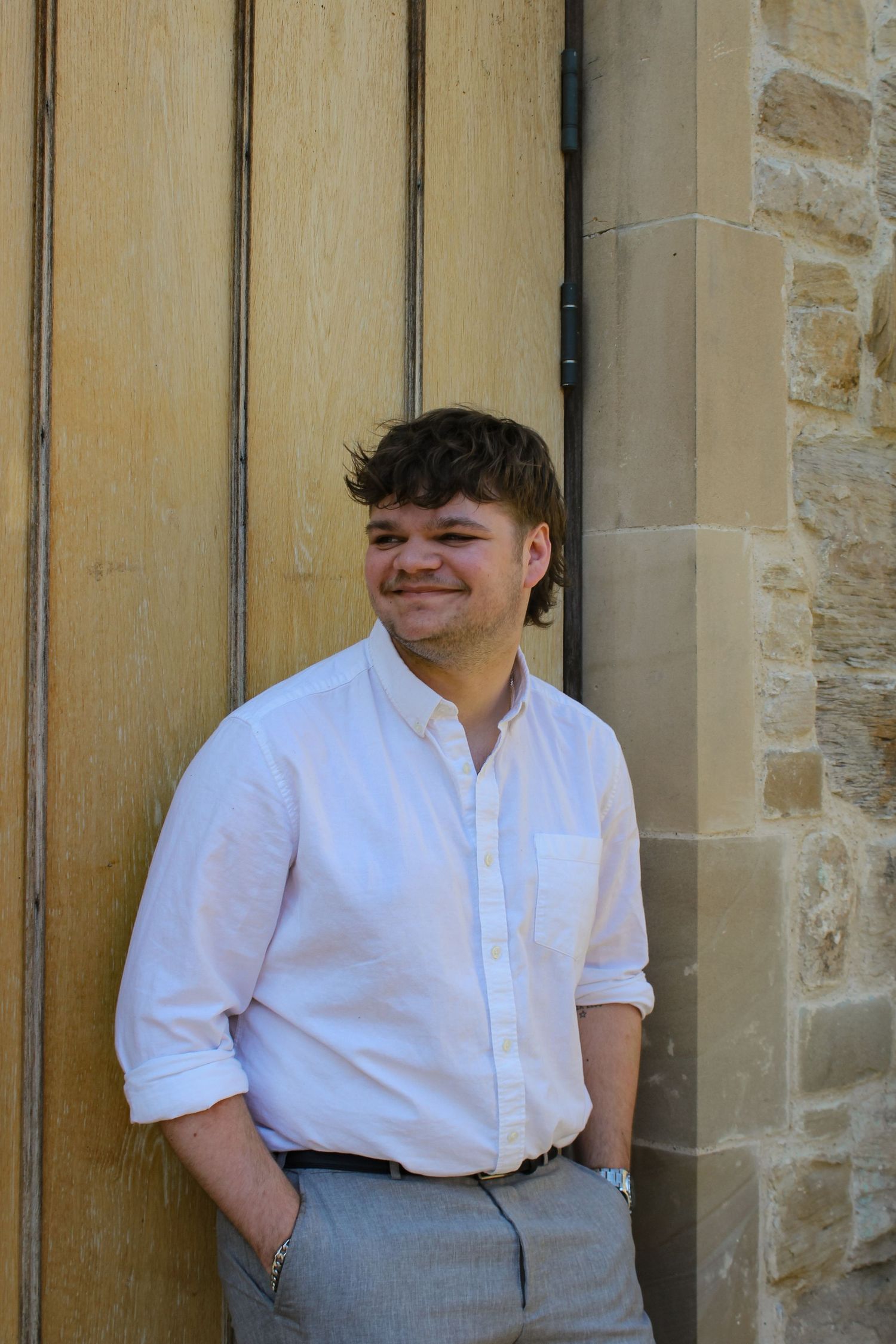 Editorial fashion portrait of a man leaning against a door. Wearing a white shirt and grey trousers with a brown belt.