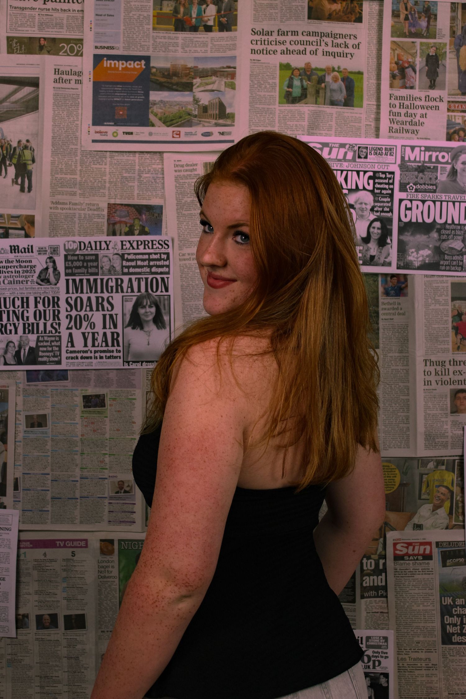 Editorial portrait of a woman smiling. The background is a wall covered with newspapers. Pink tint to the photo.