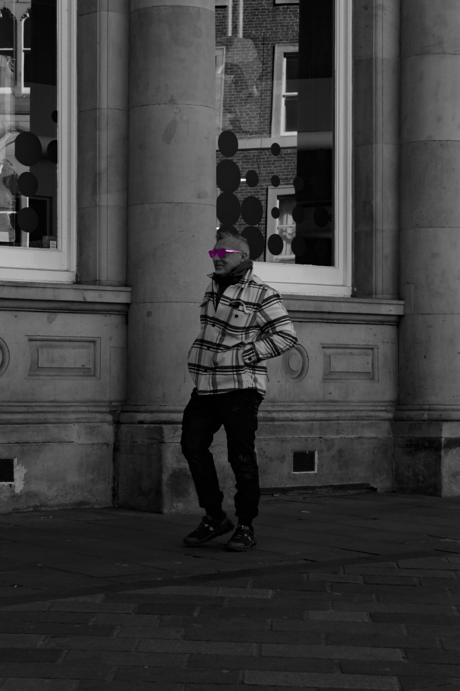 Street photography. Mostly black and white photo of a man walking down the street. His glasses are pink.