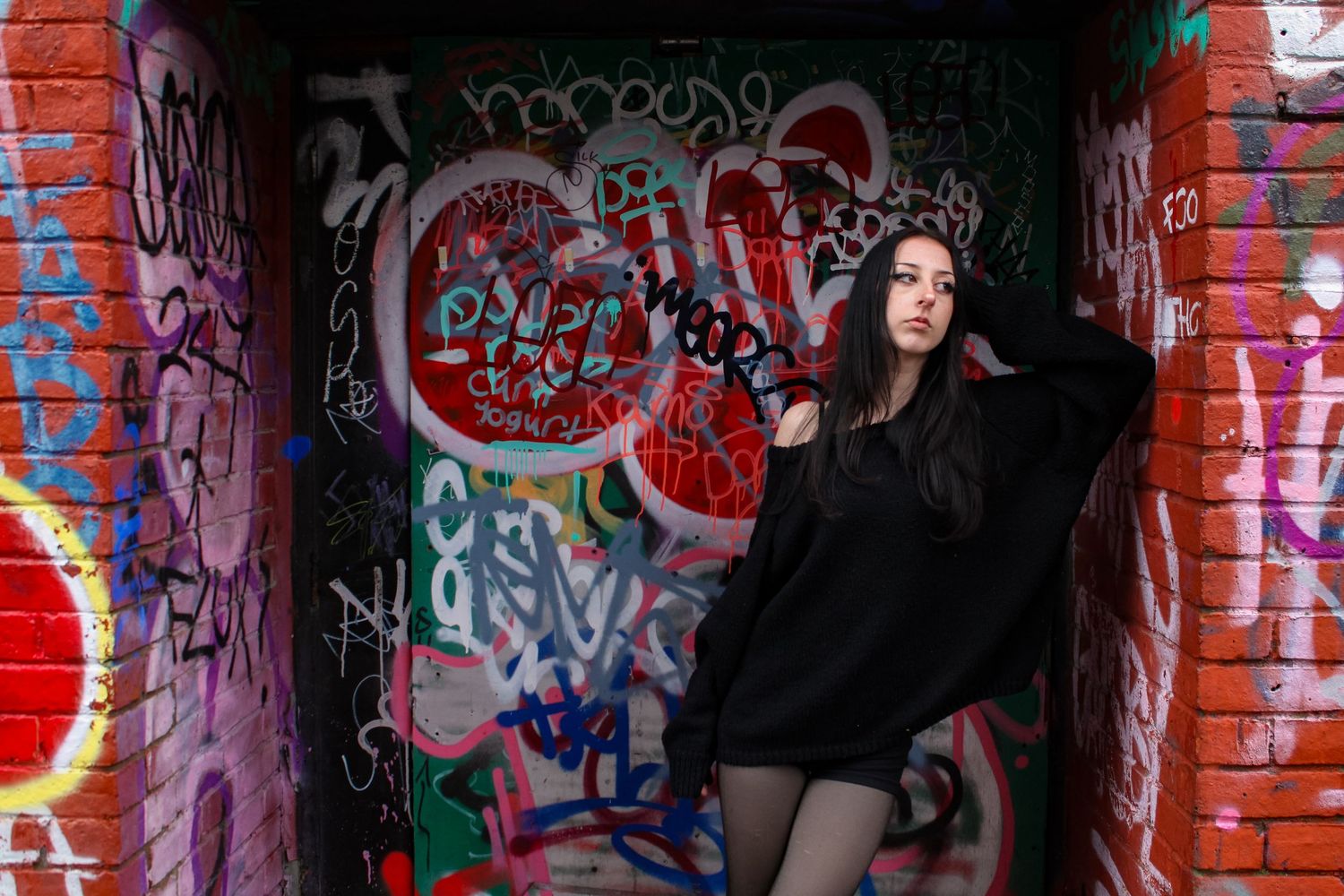 Street fashion portrait of a woman leaning on a wall. Graffiti in the background. She is wearing a black jumper, shorts and tights.