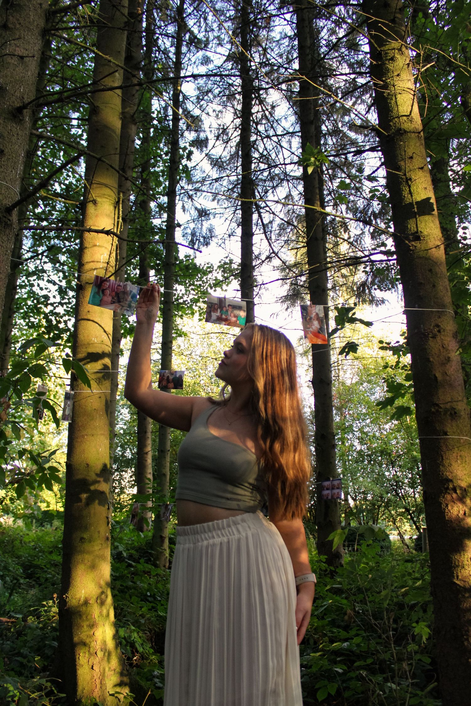 Portrait of a girl in a forest with printed photos hung up on string from tree to tree. She is looking at one of the printed photos.