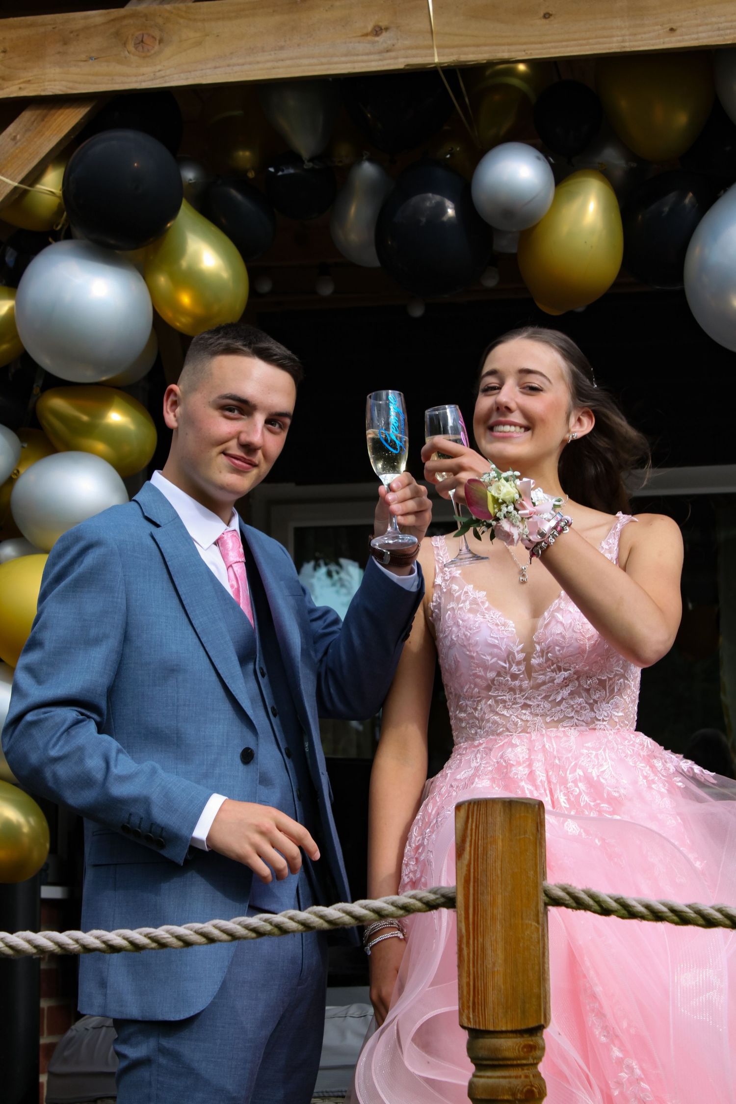 A prom photo of a couple cheers glasses. Both are smiling. His suit is blue and his tie is pink. Her dress is pink.