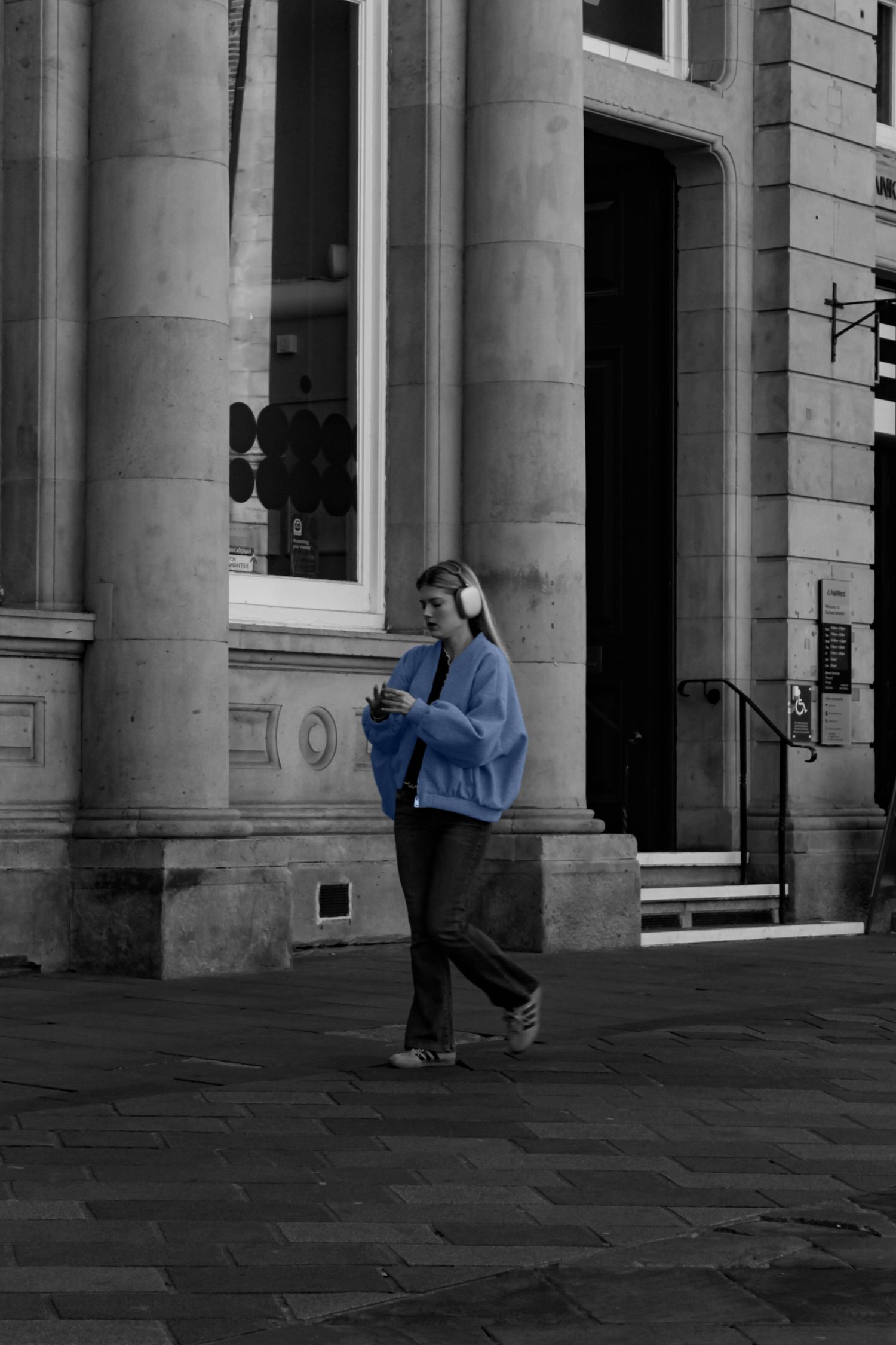 Street photography. Mostly black and white photo of a woman walking down the street. Her jacket is blue.