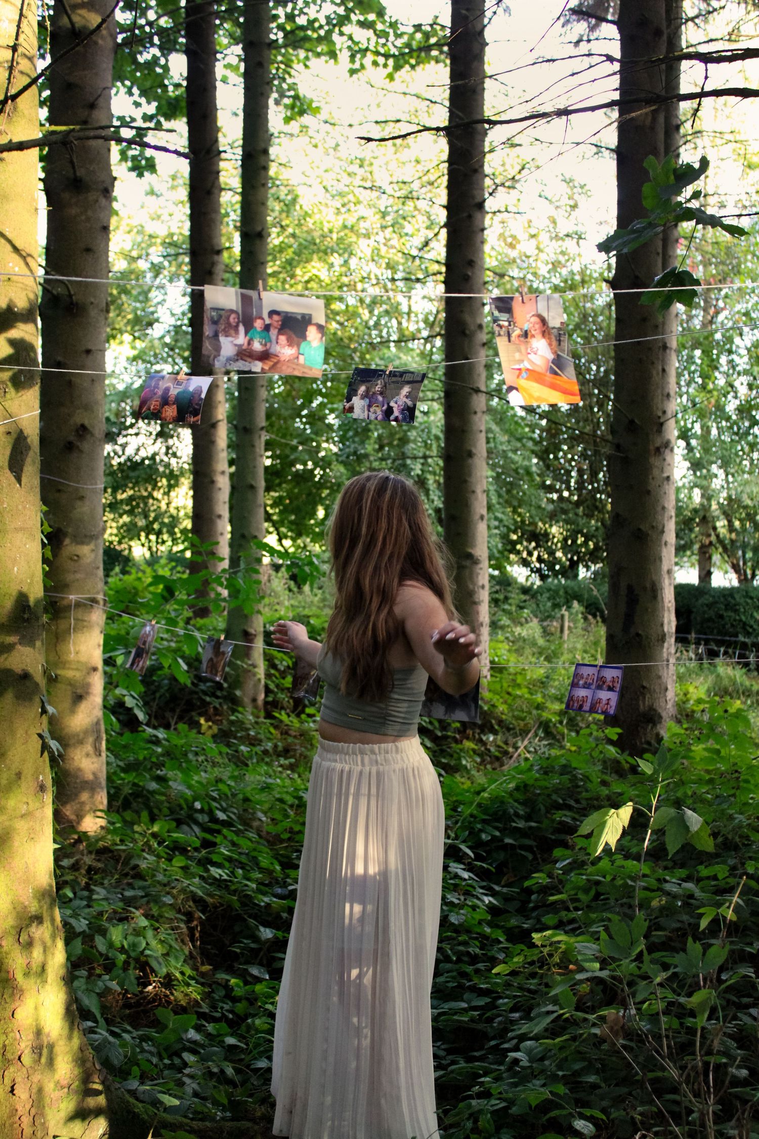 Portrait of a girl in a forest with printed photos hung up on string from tree to tree. She is dancing.