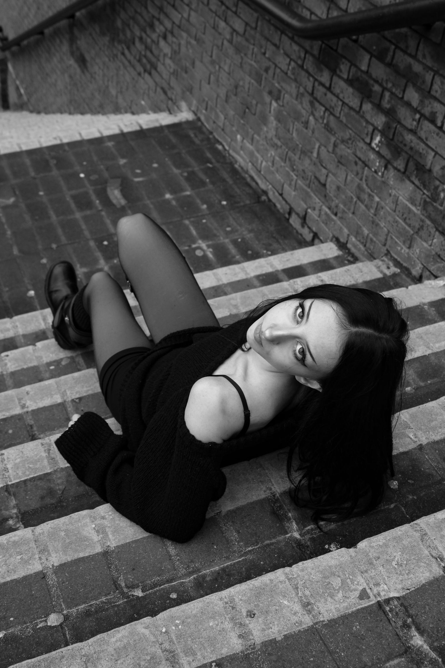 Street fashion portrait of a woman sat on a staircase posing. She is wearing a black jumper, shorts, tights and knee high boots.
B&W.