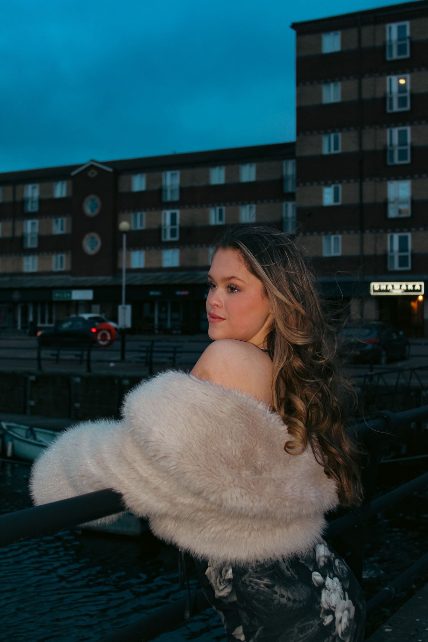 Editorial portrait photoshoot of girl wearing dress and fur coat in the evening. Looking over her shoulder pose. Flash lighting.
