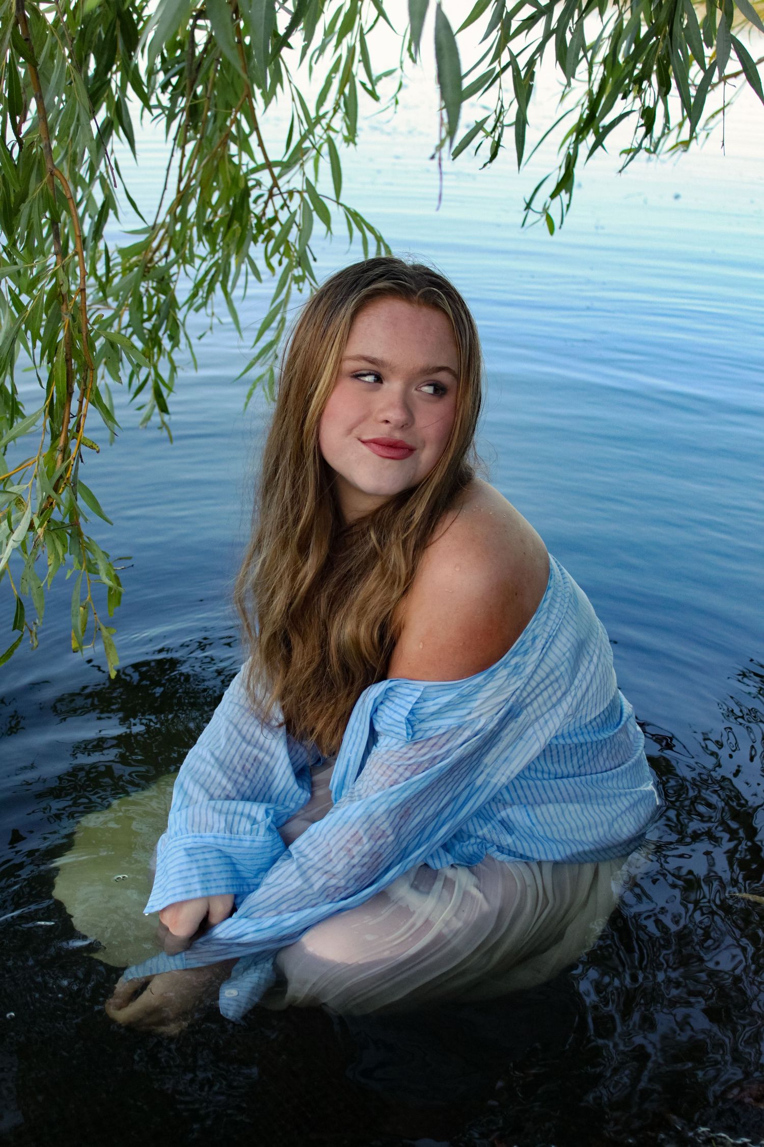 Portrait of a woman sat in a lake under a willow tree. She is wearing a blue shirt and white skirt.