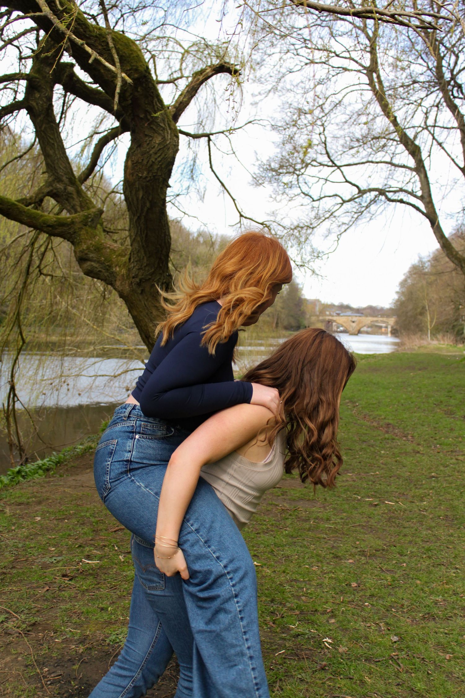 Candid photo of one girl giving another a piggyback under a willow tree. Beautiful river in the background.