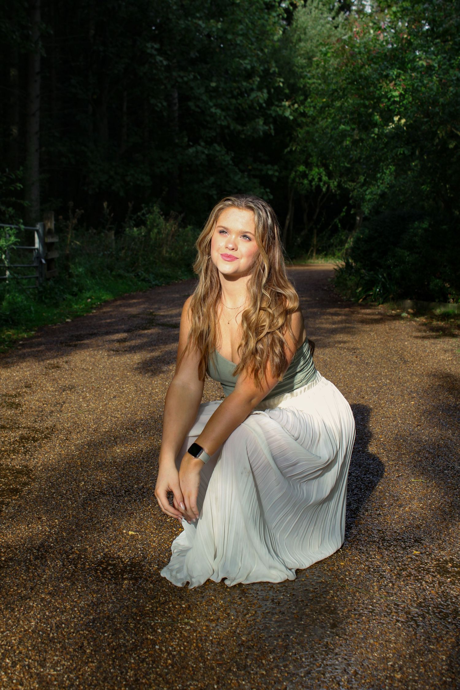 A portrait photo of a woman wearing a green top and white skirt in a park. She is in a crouched pose. Golden hour lighting.