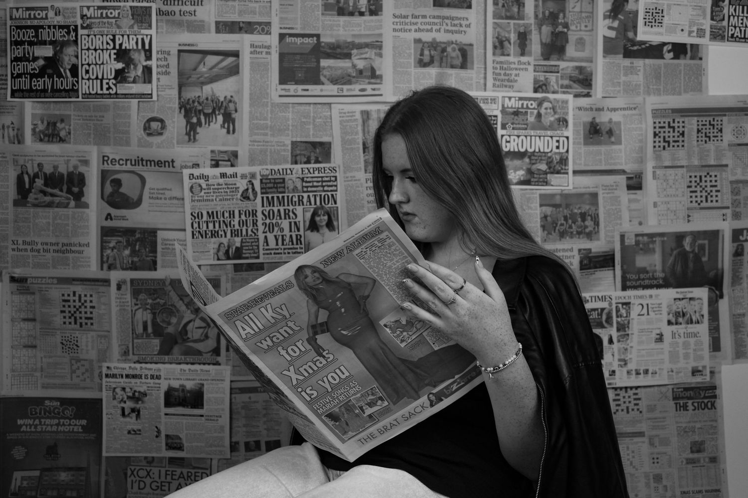 Editorial portrait of a woman sat reading a newspaper. The background is a wall covered with newspapers. Black and white.