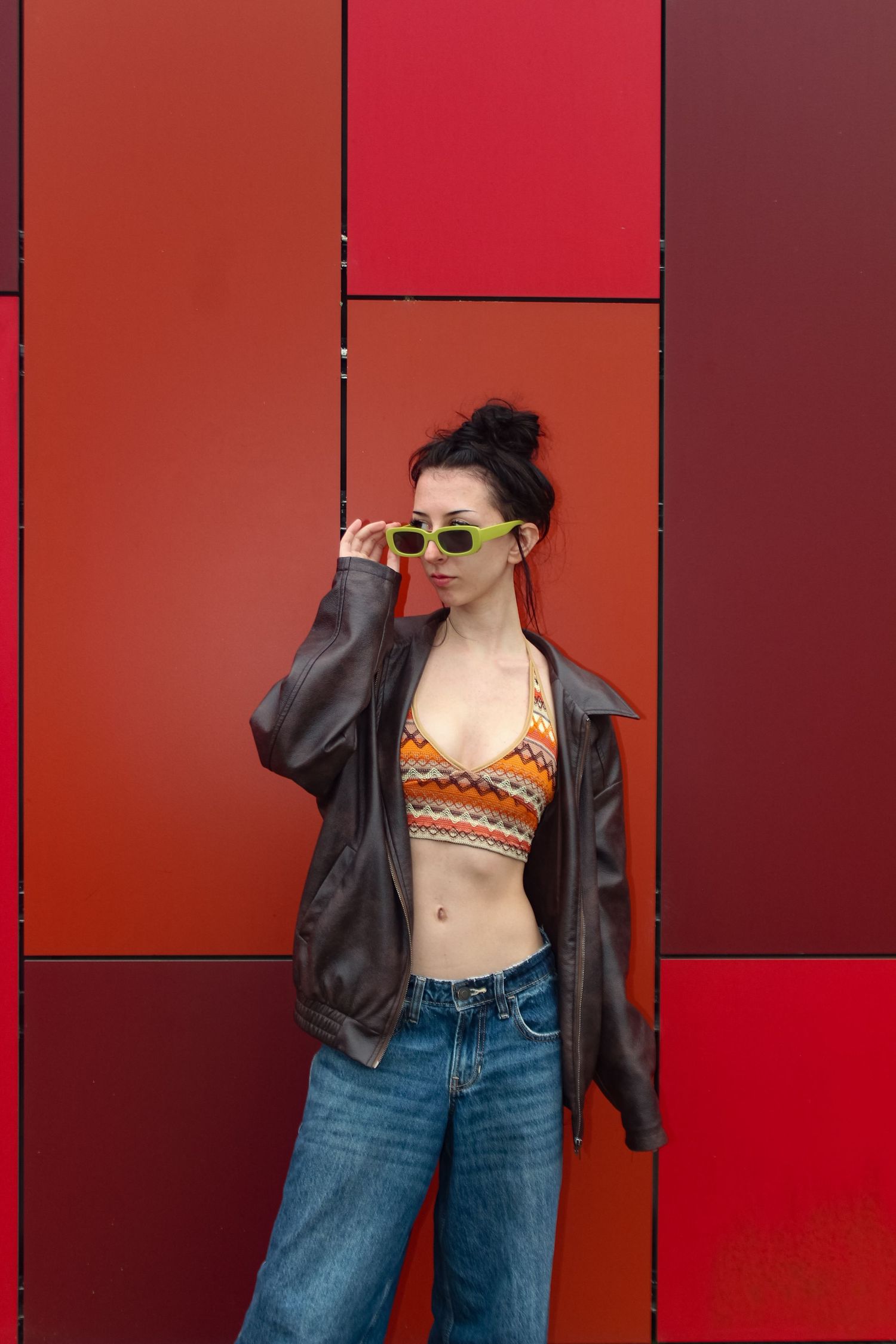 Editorial fashion portrait of a woman posed. Wearing her hair in a bun, green sunglasses, an orange top, a brown jacket and jeans.