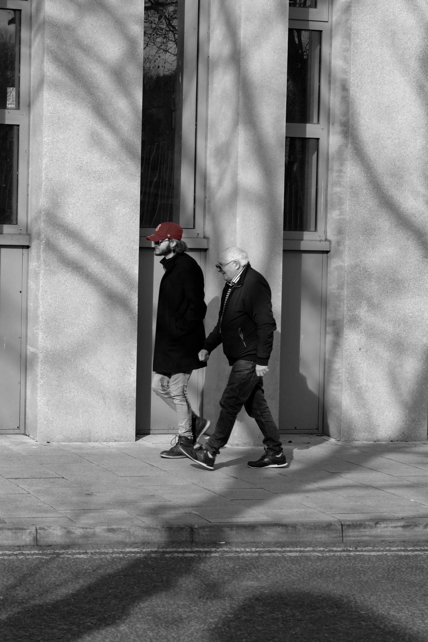 Street photography. Mostly black and white photo of two men walking down the street. One man’s hat is red.