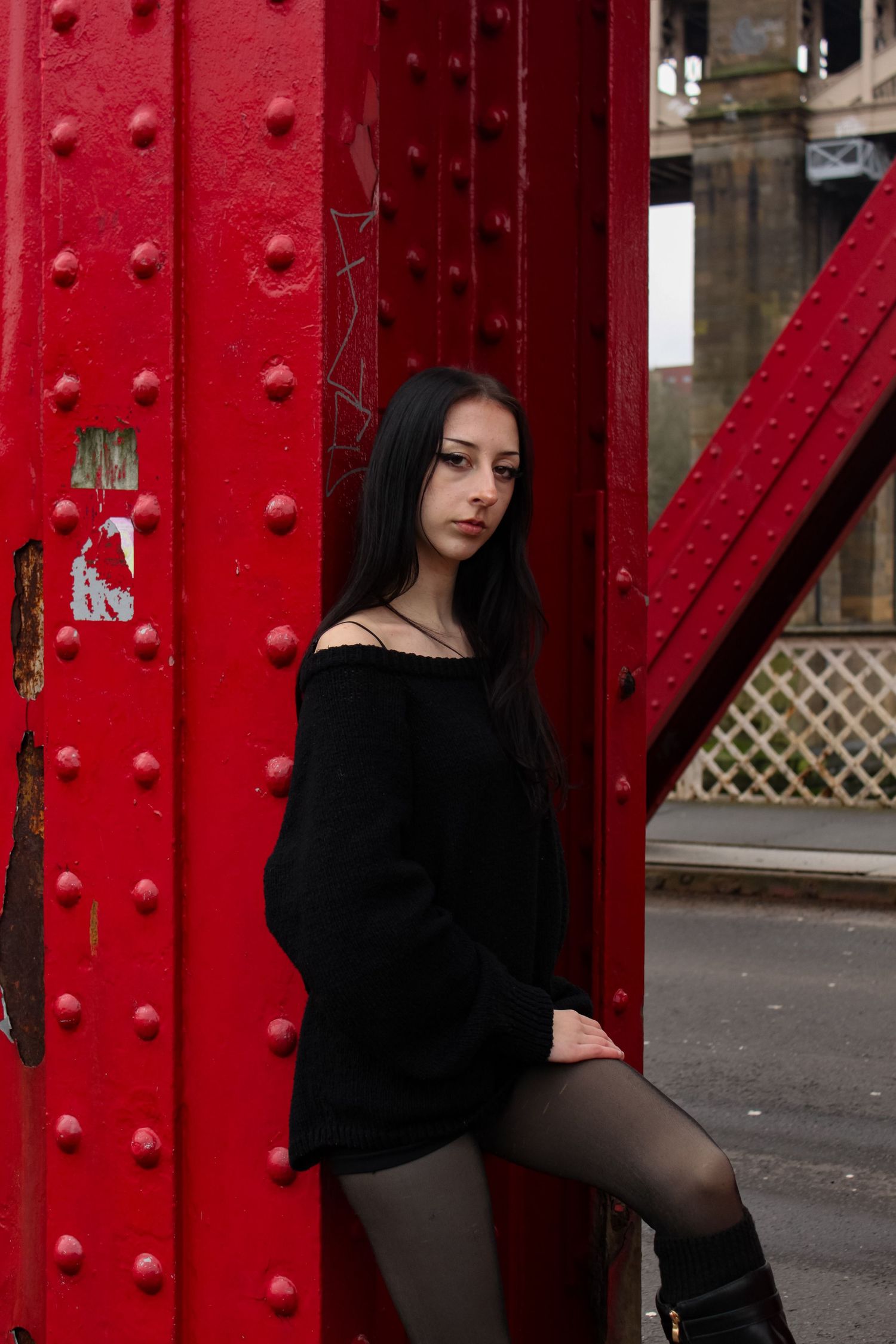 Street fashion portrait of a woman leaning on a red bridge arch. She is wearing a black jumper, shorts and knee high boots. Newcastle.