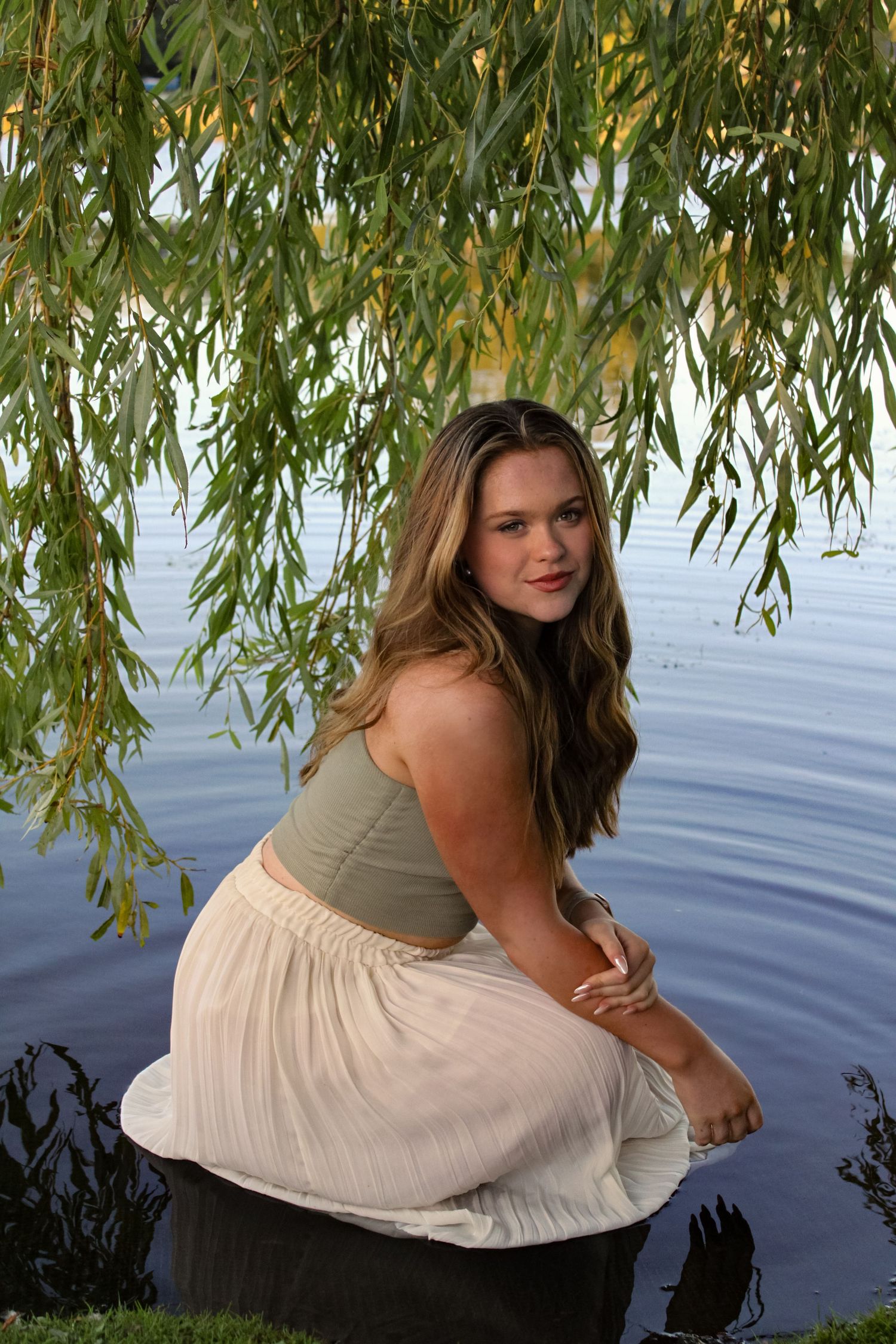 Portrait of a woman sat in a lake under a willow tree. She is wearing a green top and white skirt.