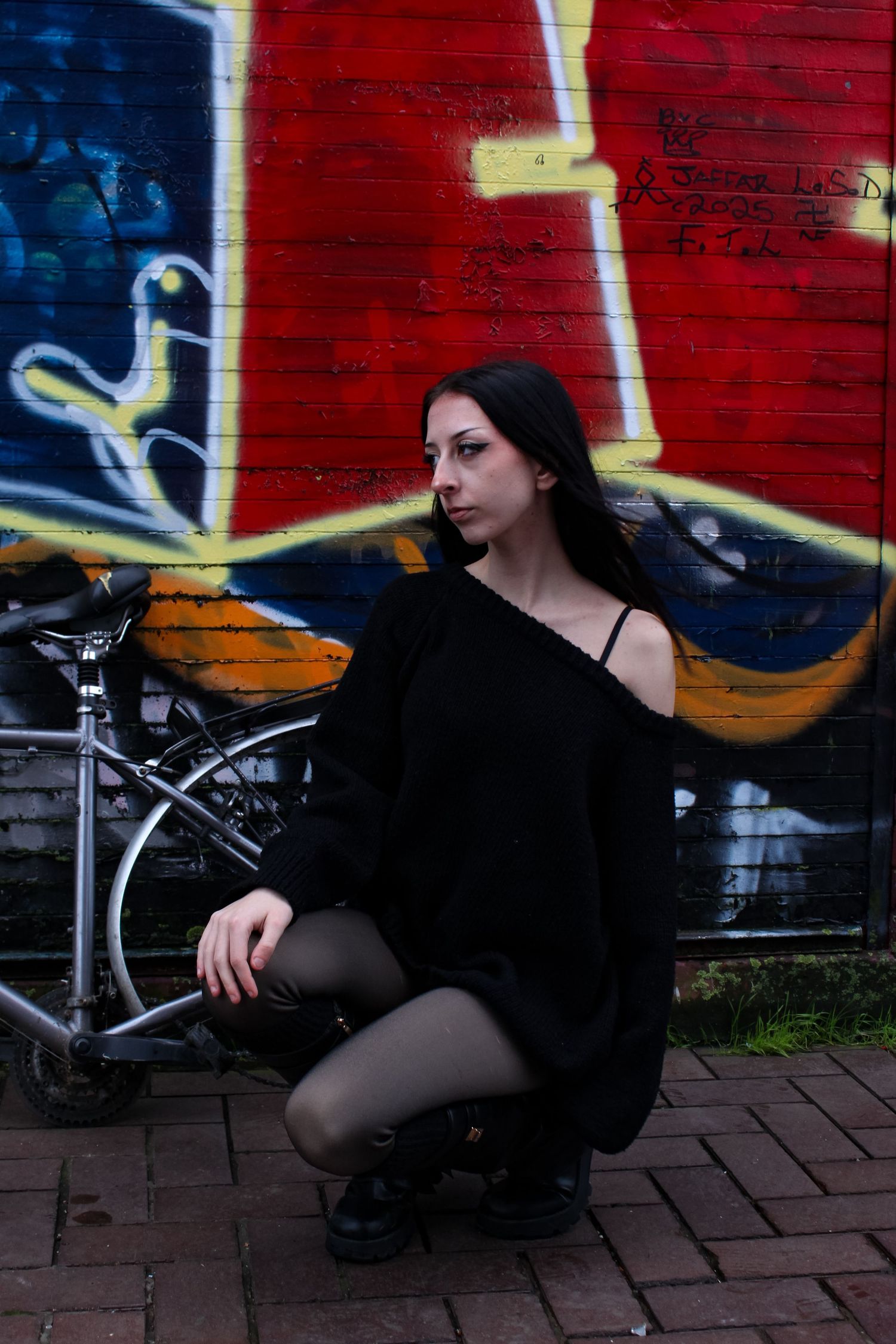 Street fashion portrait of a woman in a crouched pose. Graffiti and an abandoned bike in the background. She is wearing a black jumper.