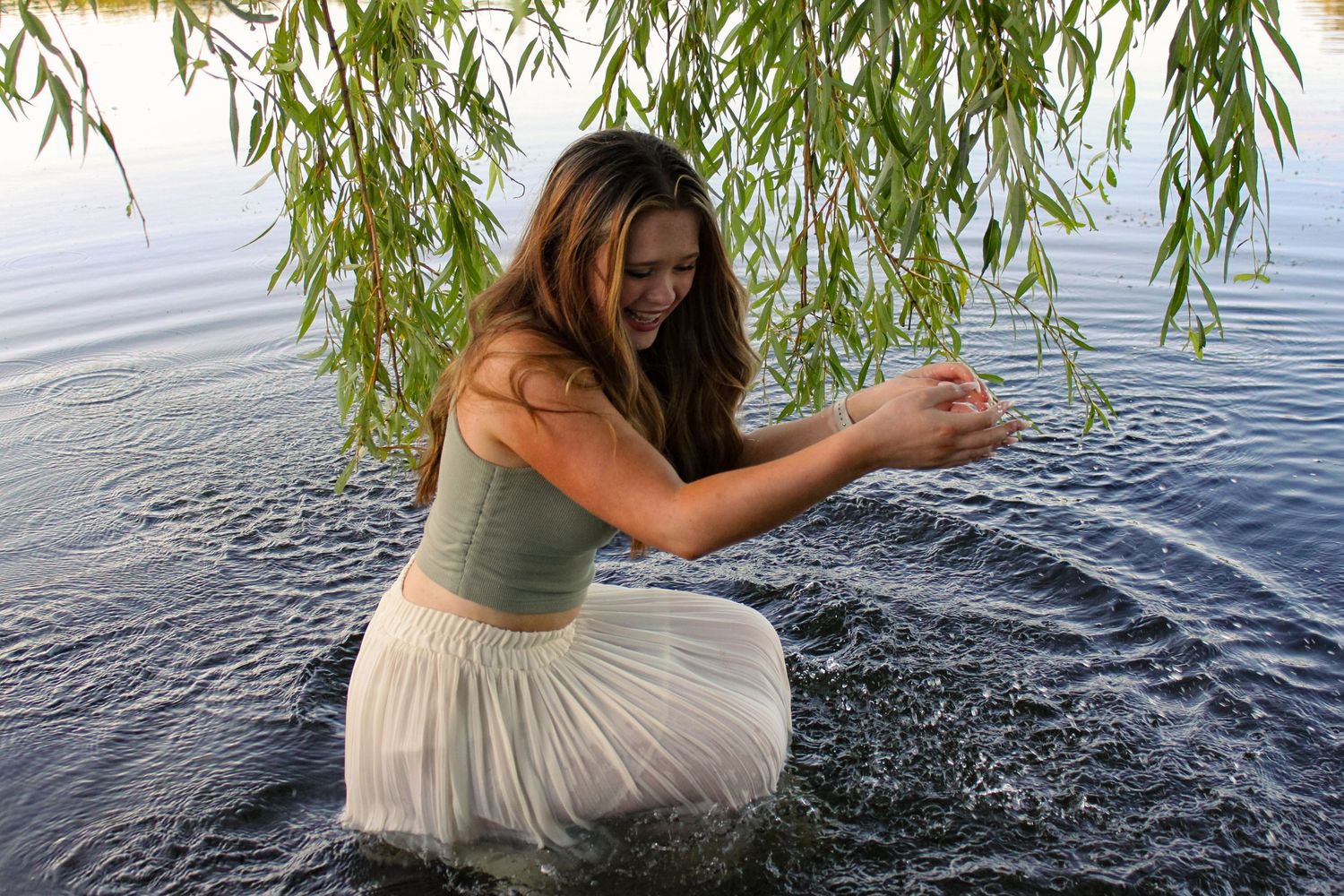 Portrait of a woman sat in a lake under a willow tree and playing with the water. She is wearing a green top and white skirt.
