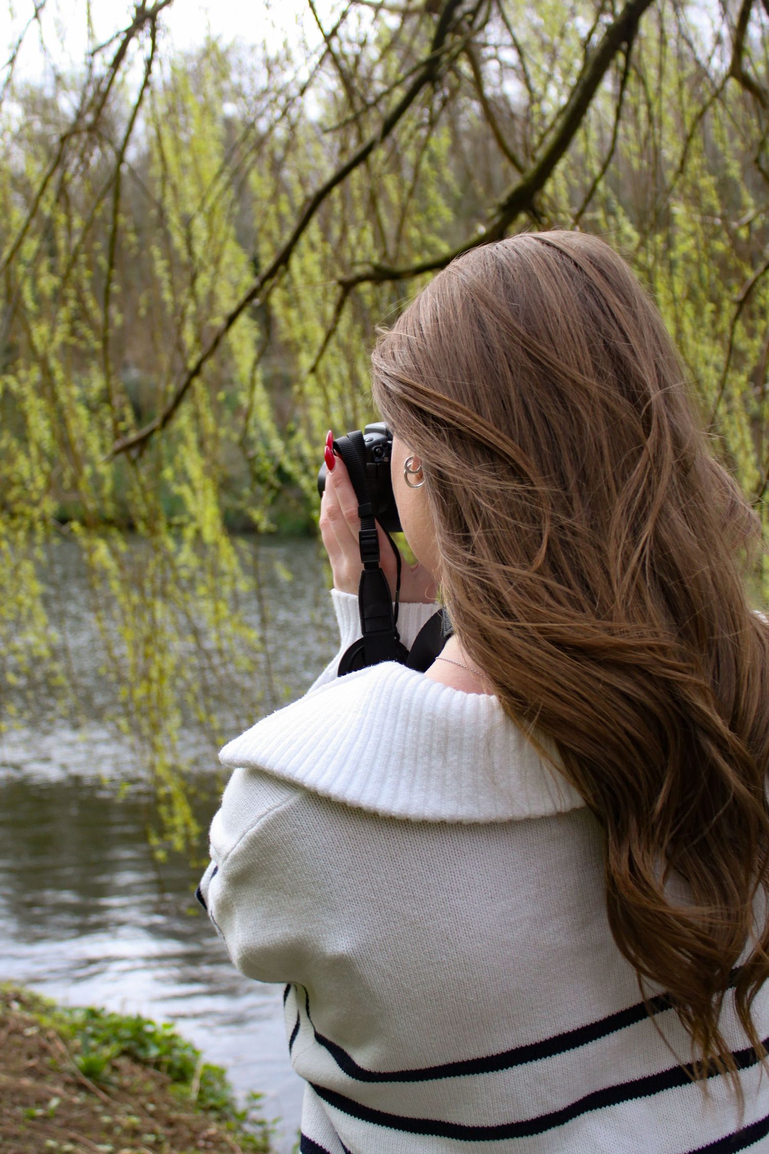 A portrait of photographer Ella Nicole taking a photo with her camera under a willow tree. River in the background.