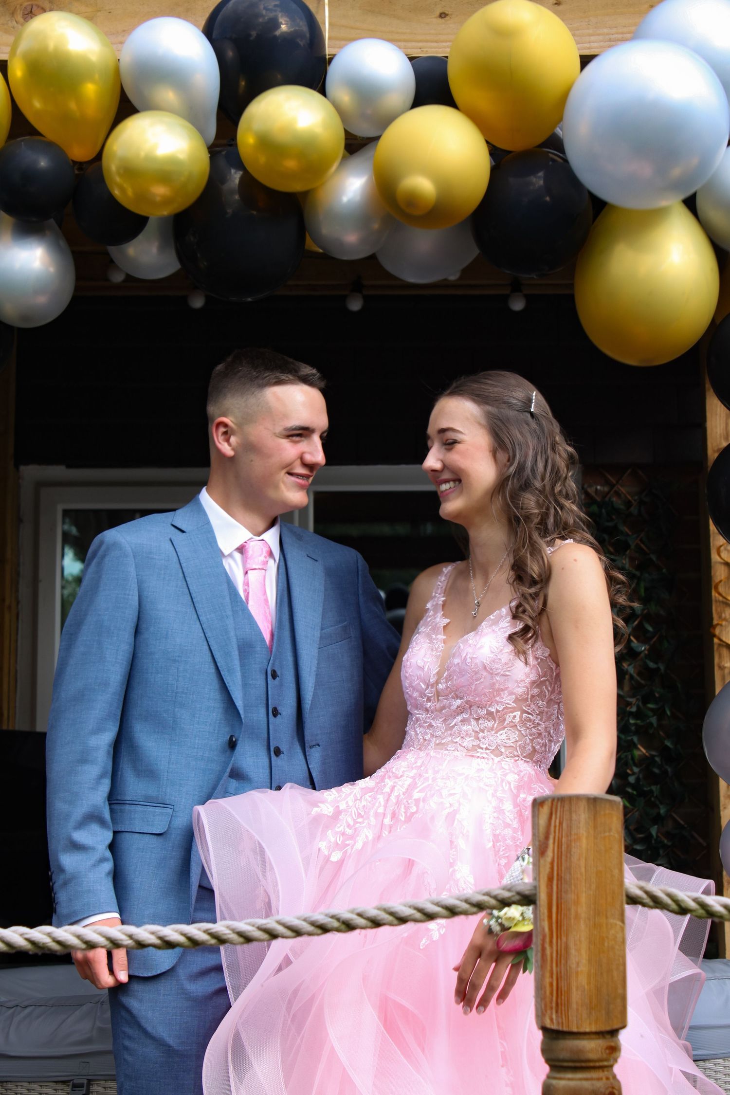 A couple on their prom day laughing. His suit is blue and his tie is pink. Her prom dress is pink.