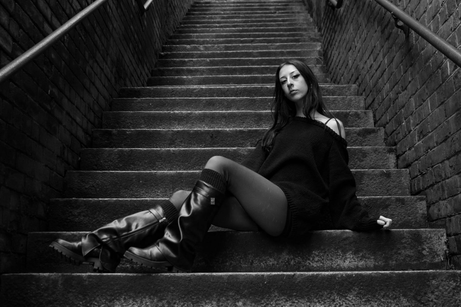 Street fashion portrait of a woman sat on a staircase posing. She is wearing a black jumper, shorts, tights and knee high boots. B&W.