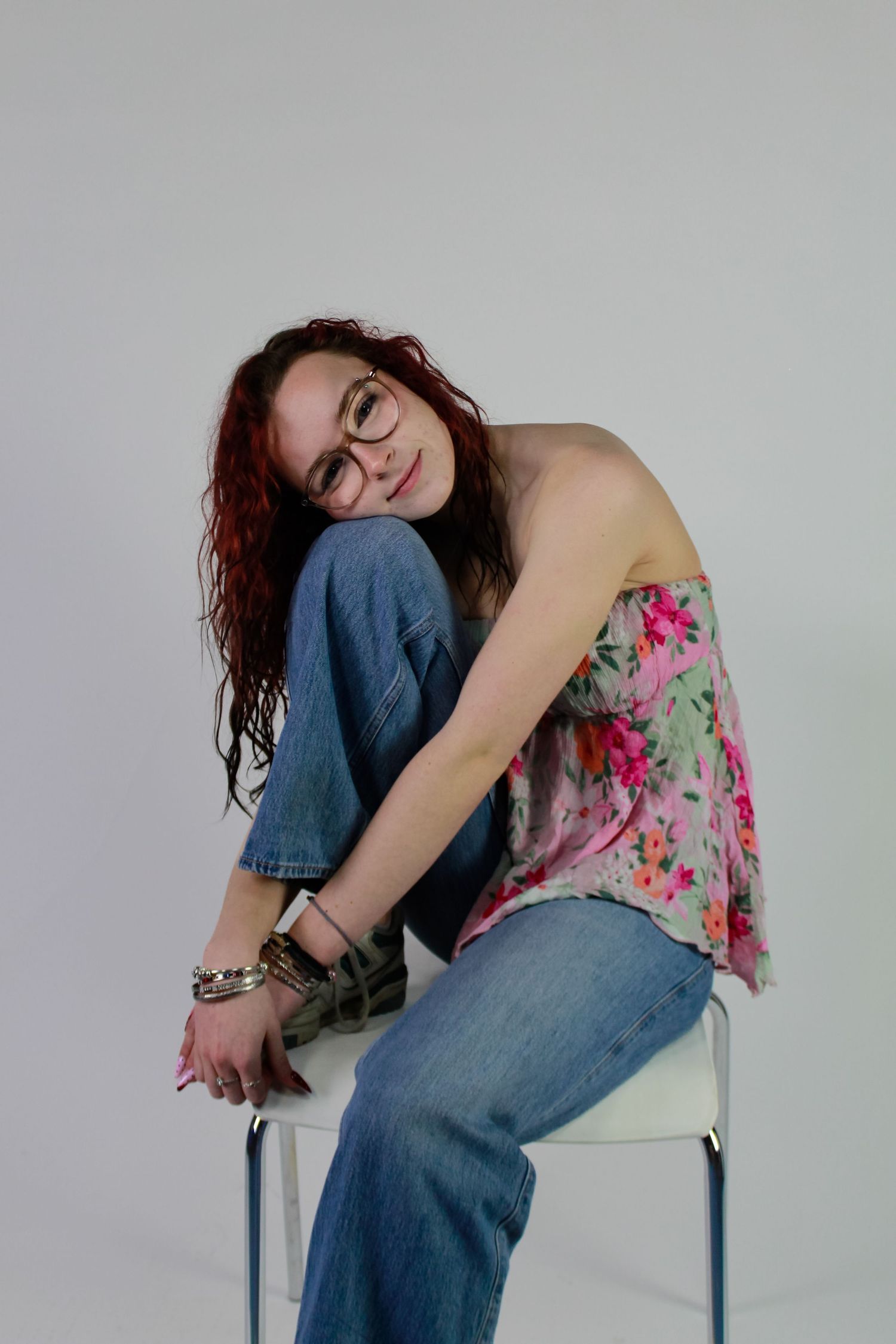 Studio portrait of a woman posed sat causally on a chair. Fashion photo. Wearing her red hair down, jeans and pink floral top.