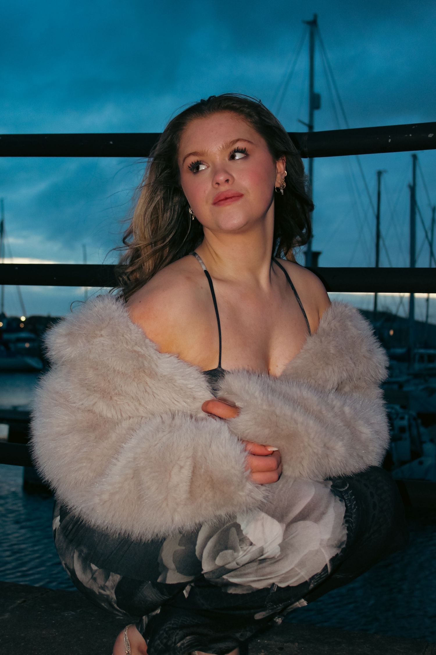 Editorial portrait photoshoot of girl wearing dress and fur coat in the evening. Crouched pose. Flash lighting.
