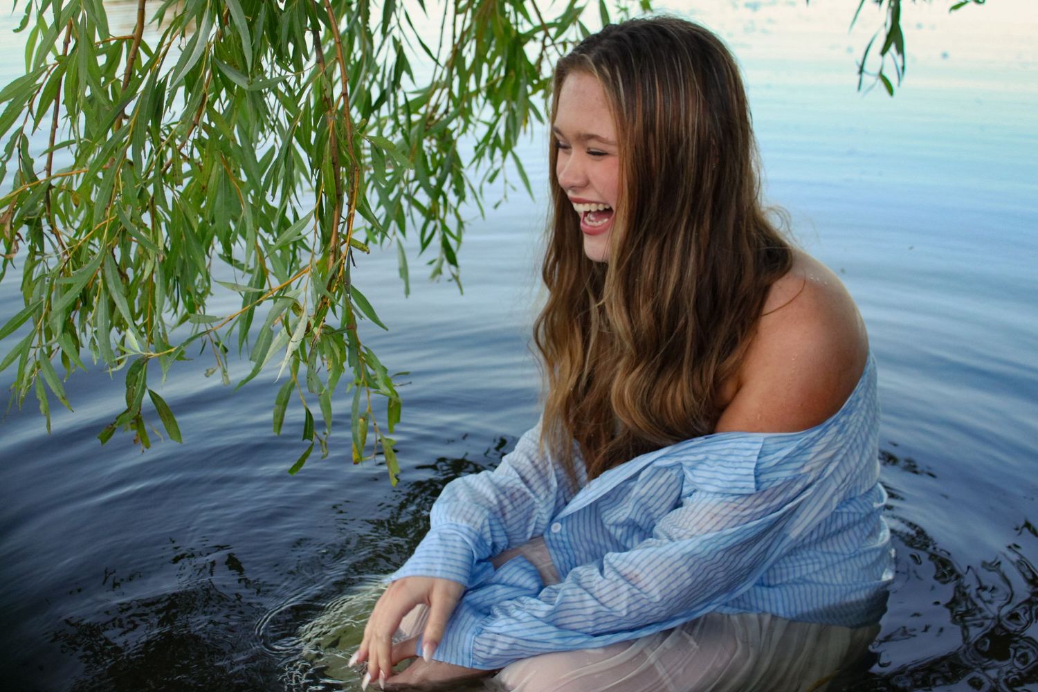 Portrait of a woman sat in a lake under a willow tree. She is wearing a blue shirt and white skirt.
