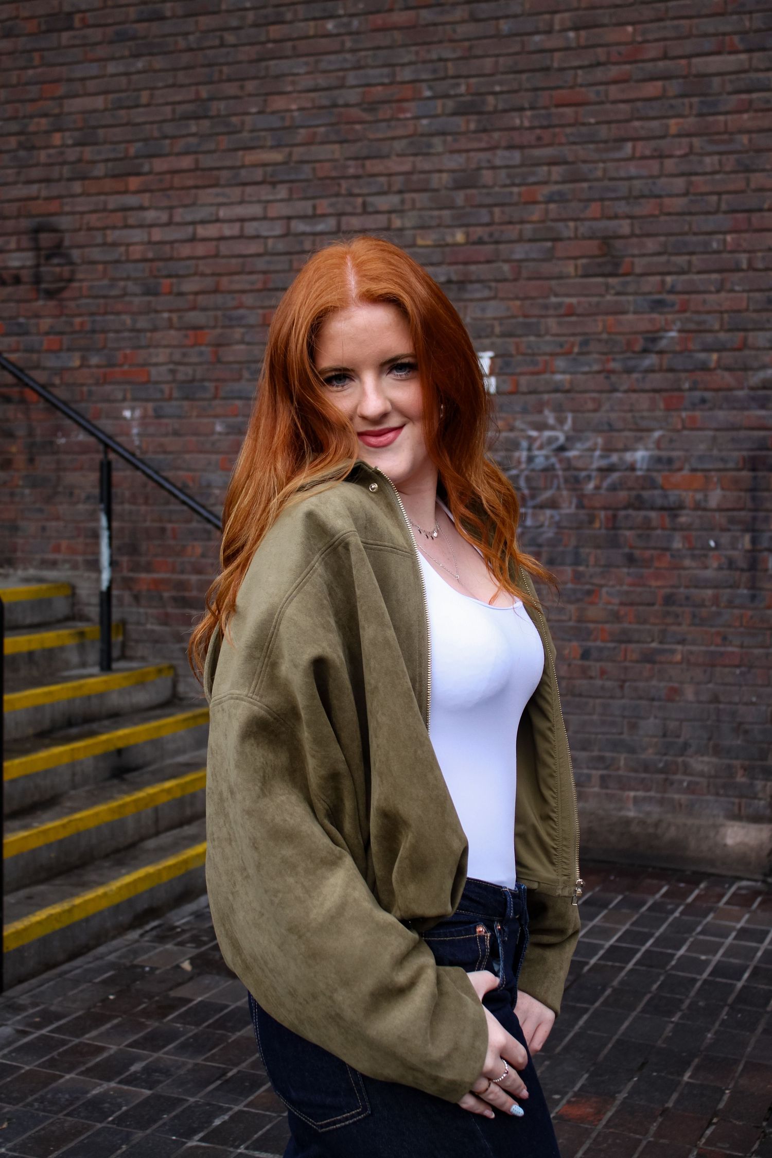 Street fashion portrait photography of a woman smiling. She is wearing jeans, a white top and a green jacket.
