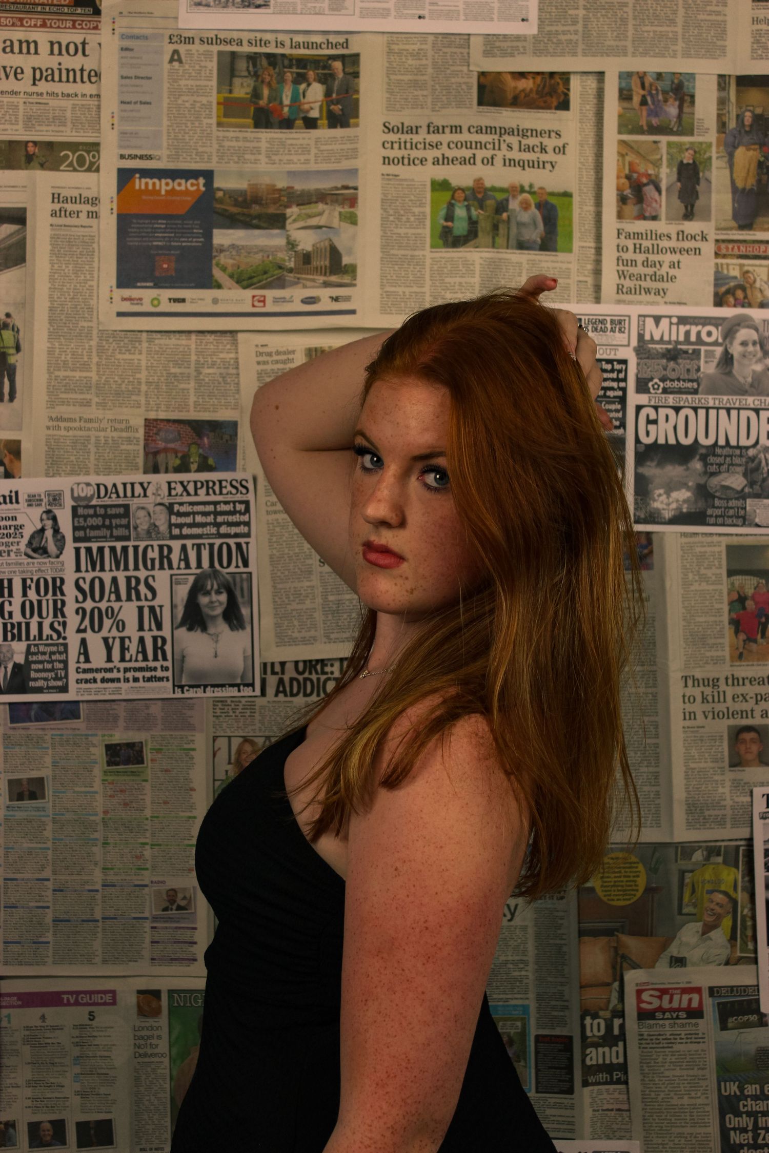 Editorial portrait of a woman posing. The background is a wall covered with newspapers. Pink tint to the photo.