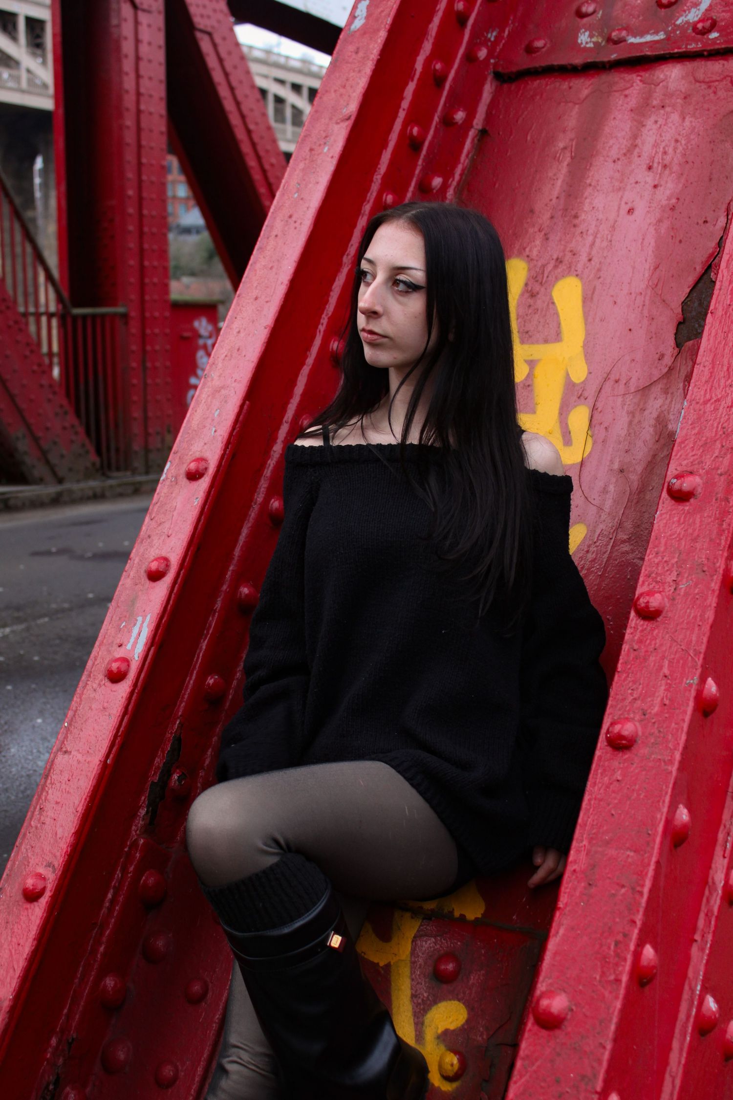 Street fashion portrait of a woman leaning on a red bridge arch. She is wearing a black jumper, shorts and knee high boots. Newcastle.
