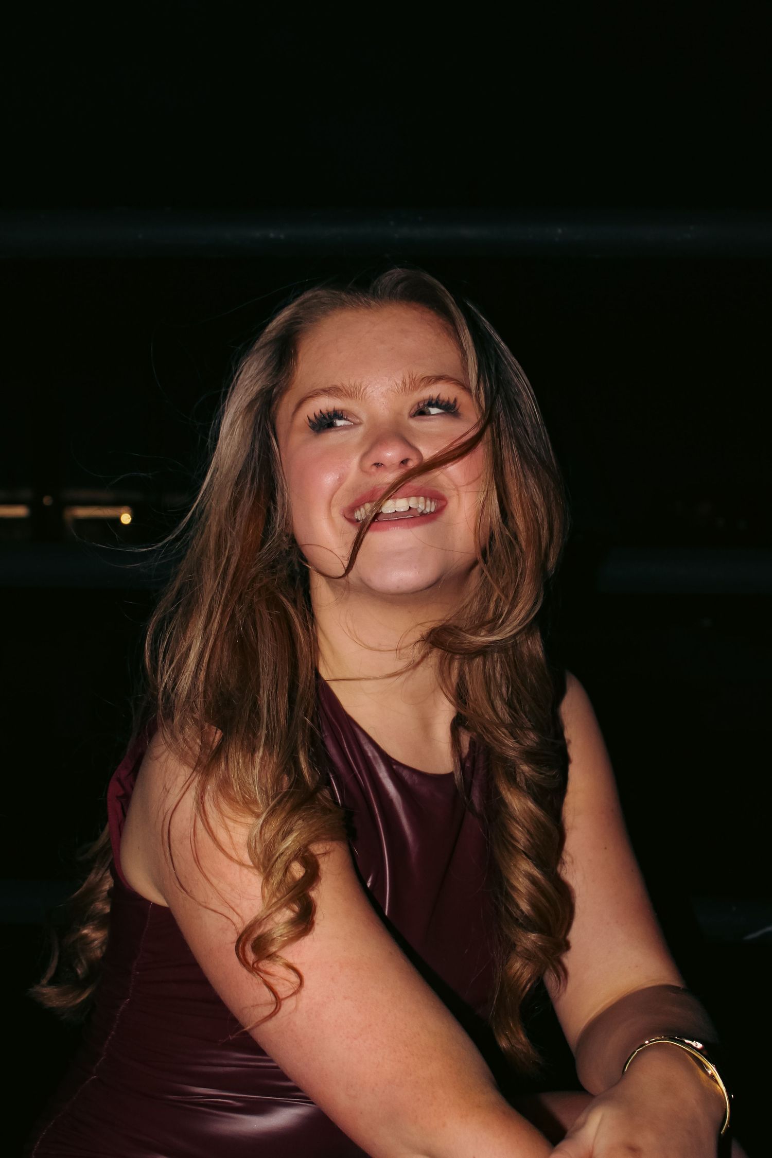 Editorial portrait photoshoot of girl laughing and wearing burgundy dress at night. Crouched pose. Flash lighting.
