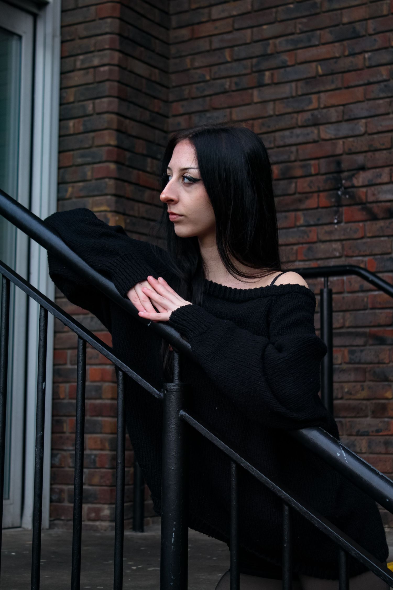 Street fashion portrait of a woman leaving on a handrail. She is looking off to the side. She is wearing a black jumper, skirt and boots.