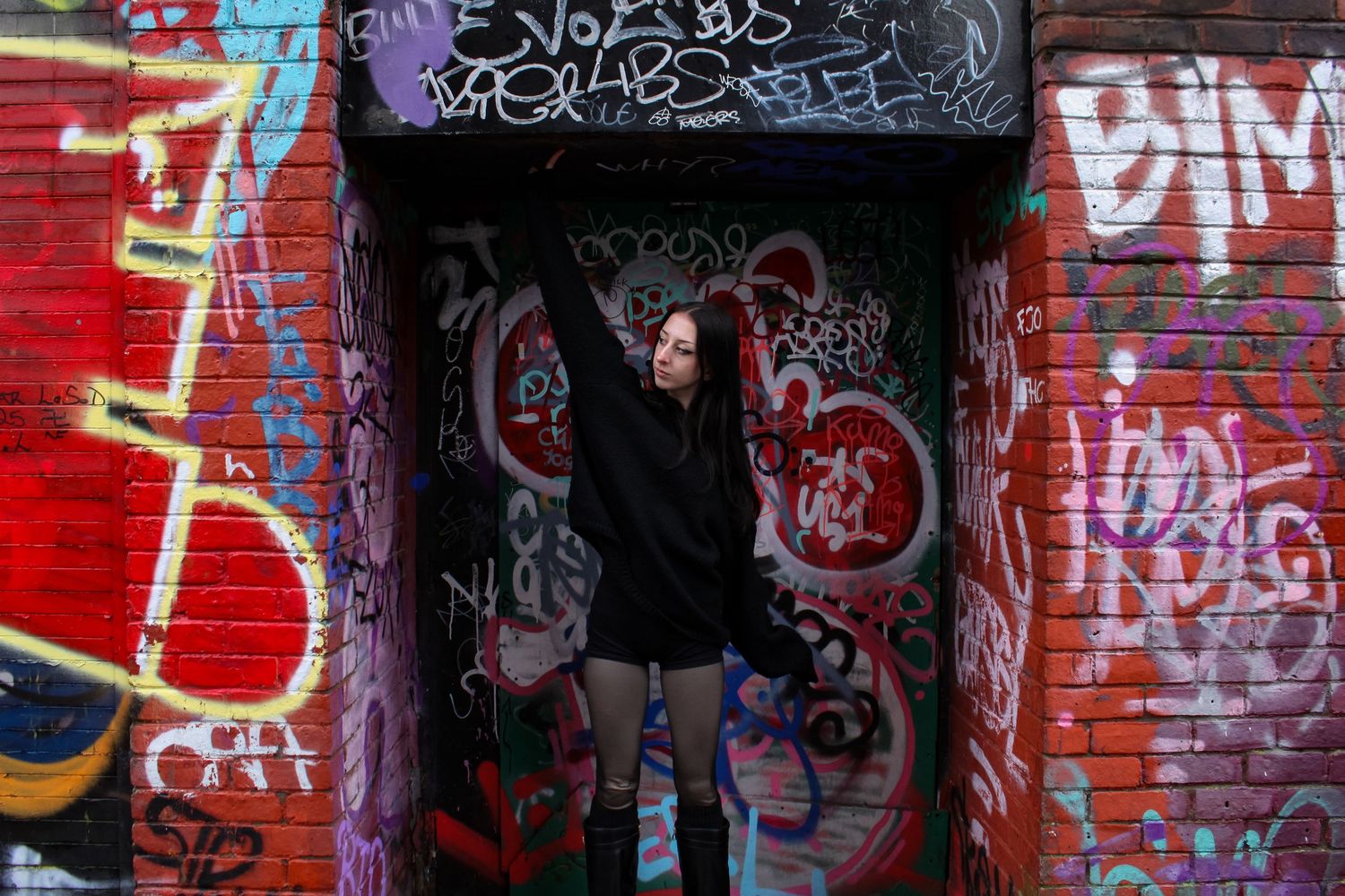 Street fashion portrait of a woman posing. Graffiti in the background. She is wearing a black jumper, shorts and knee high boots.