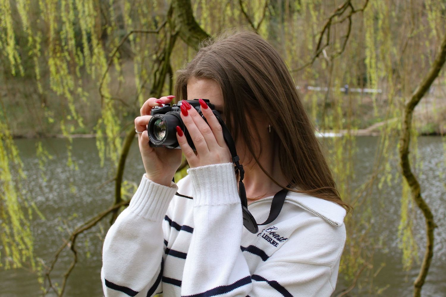 A portrait of photographer Ella Nicole taking a photo with her camera under a willow tree. River in the background.