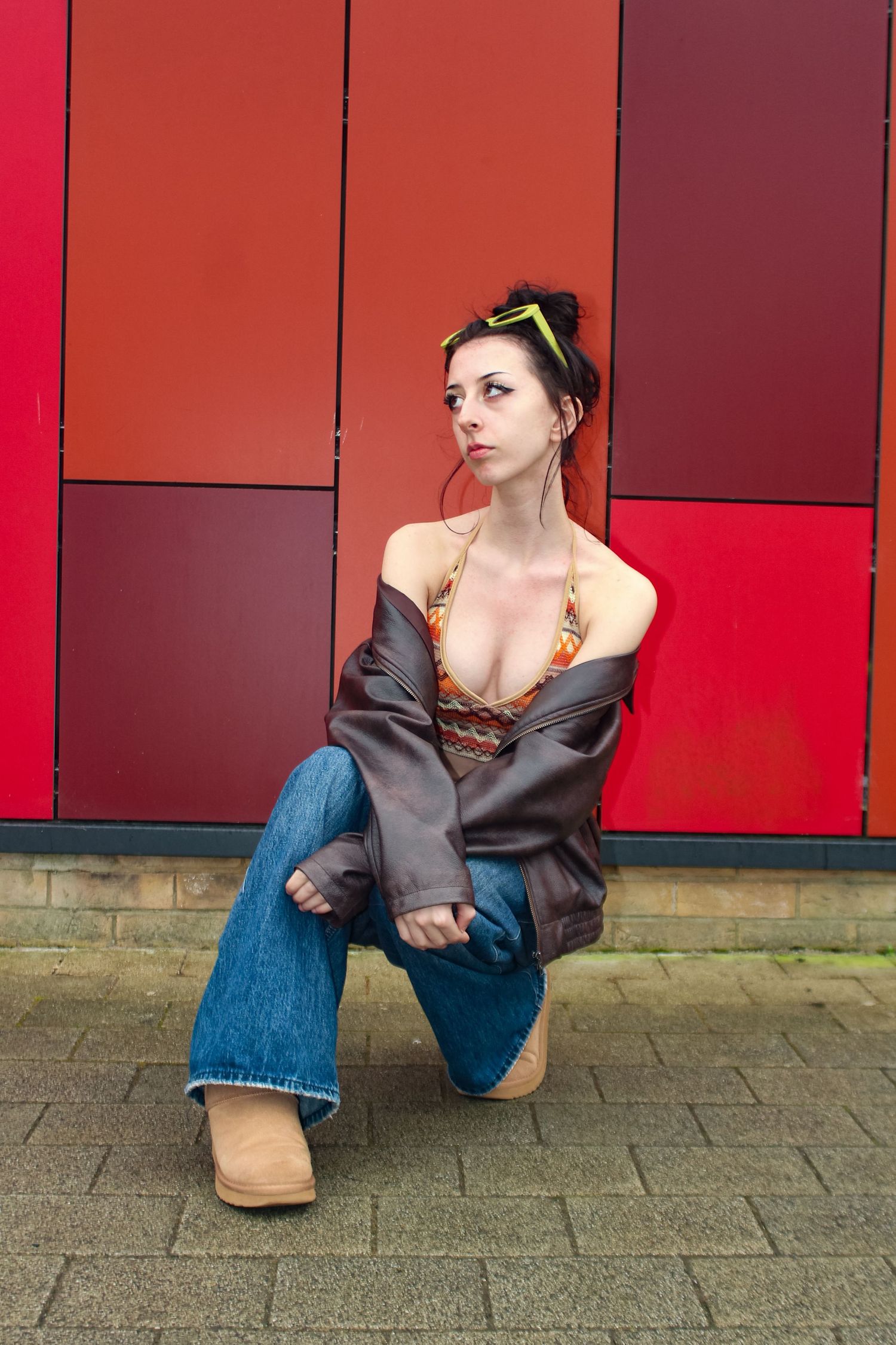 Editorial fashion portrait of a woman crouching. Wearing her hair in a bun, green sunglasses, an orange top, a brown jacket and jeans.