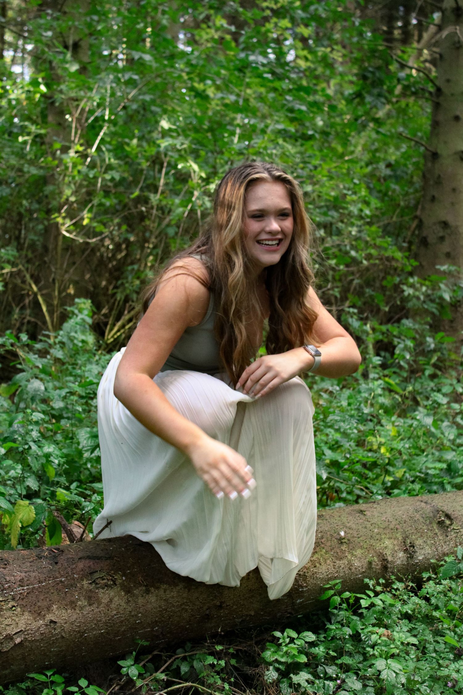 A photo of a woman crouched on a log and laughing in a forest. She is wearing a green top and white skirt.