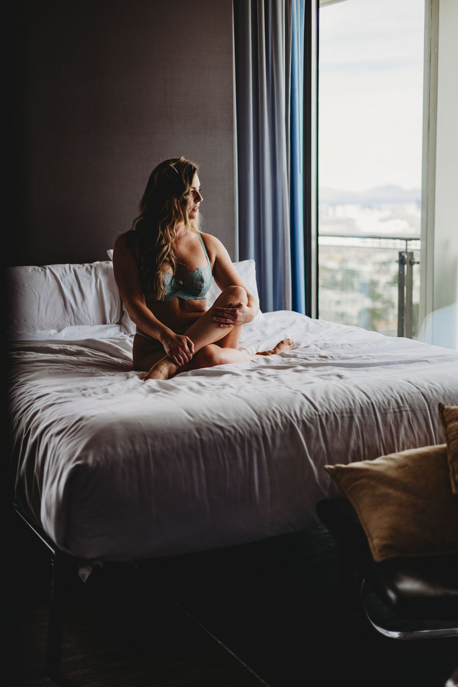 A person in casual attire sits on a bed near a window in a dimly lit room with natural light filtering through.