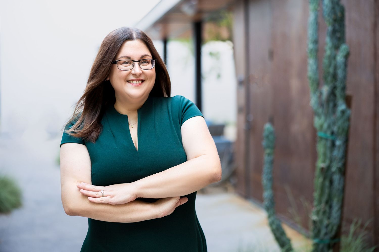 Woman in green dress with arms crossed smiling outdoors near cactus and wooden fence.