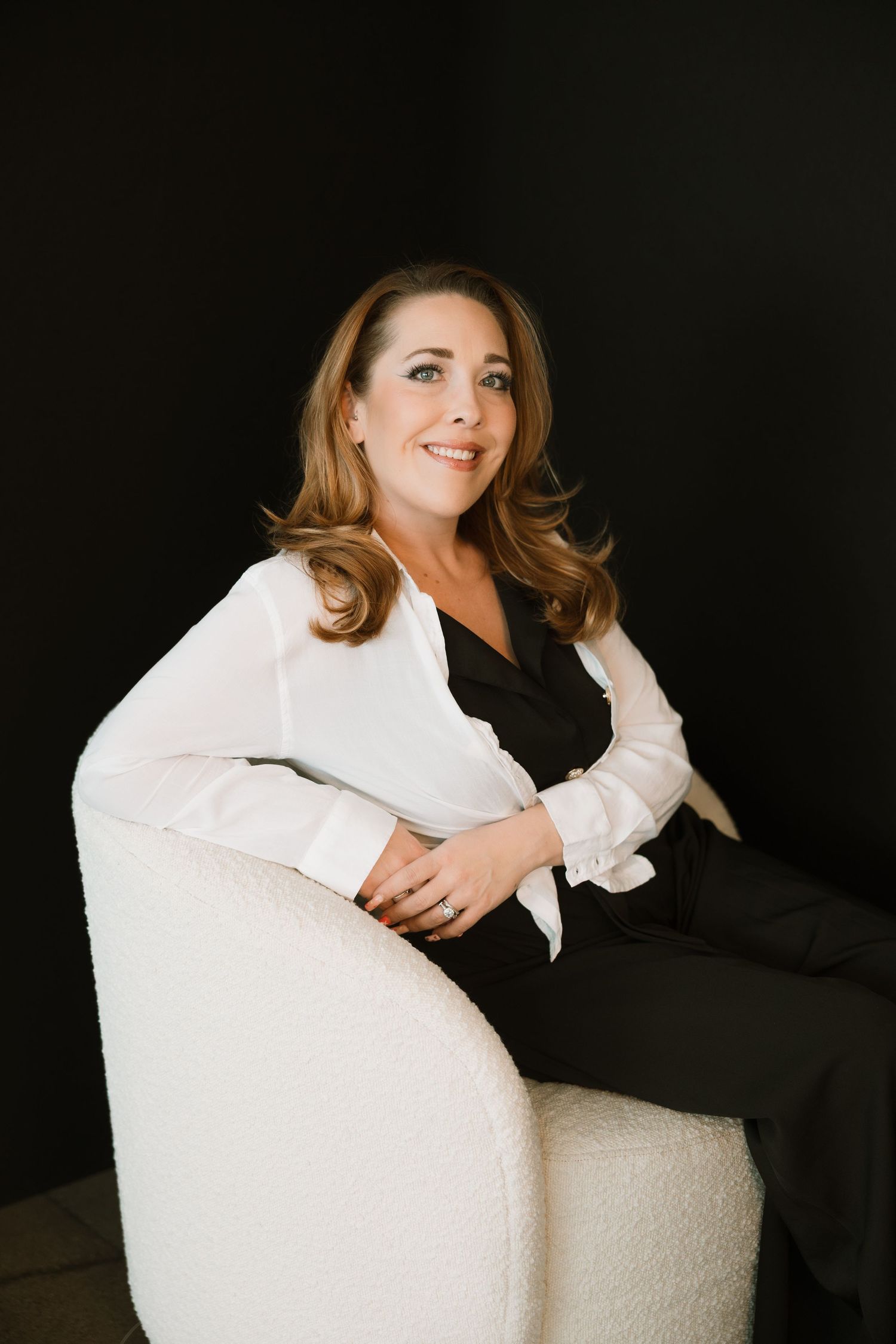 Woman in white blazer and black outfit seated in a modern white chair against a dark background.