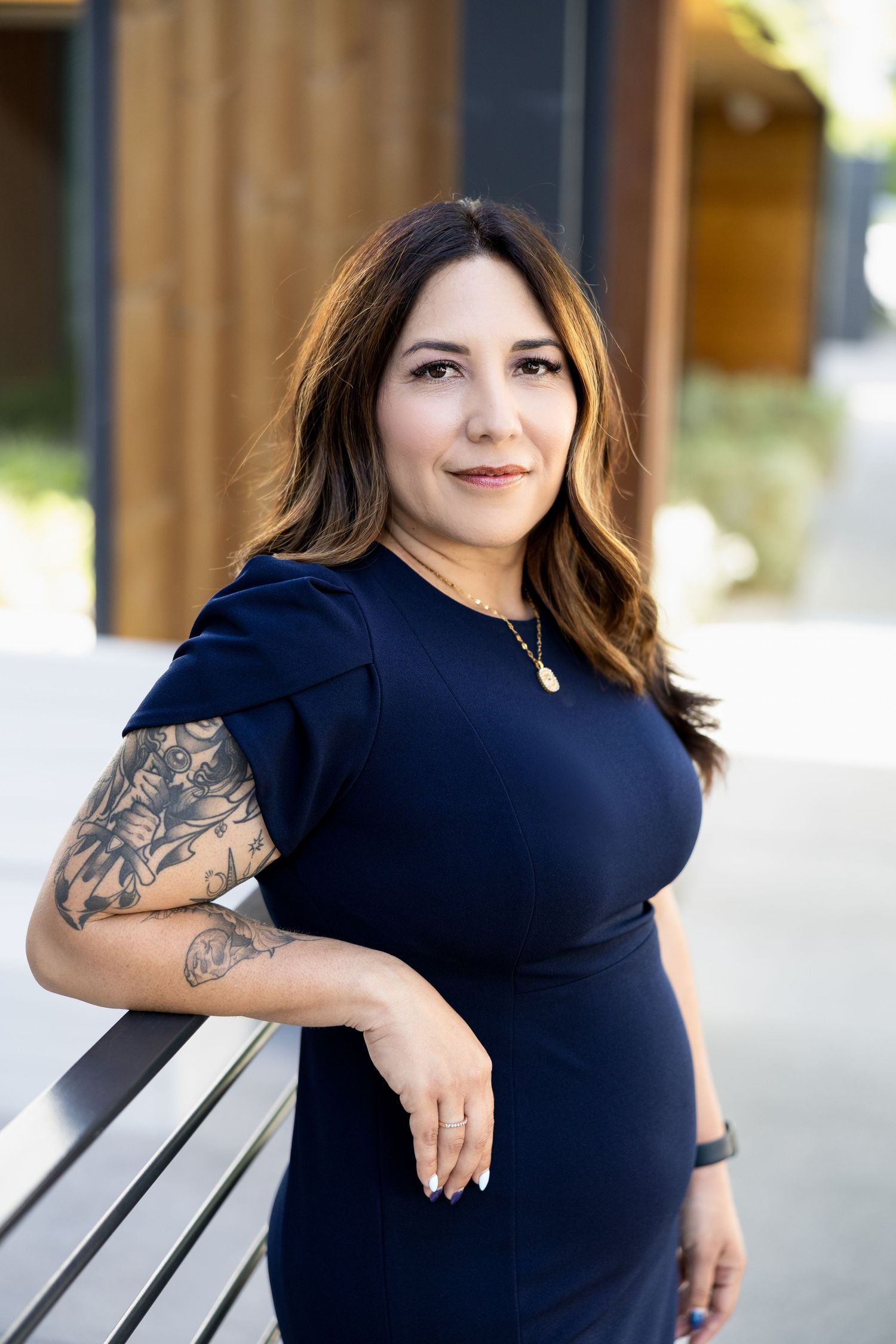 Woman in navy dress with arm tattoo leans on railing, posing outdoors in a professional portrait photo.