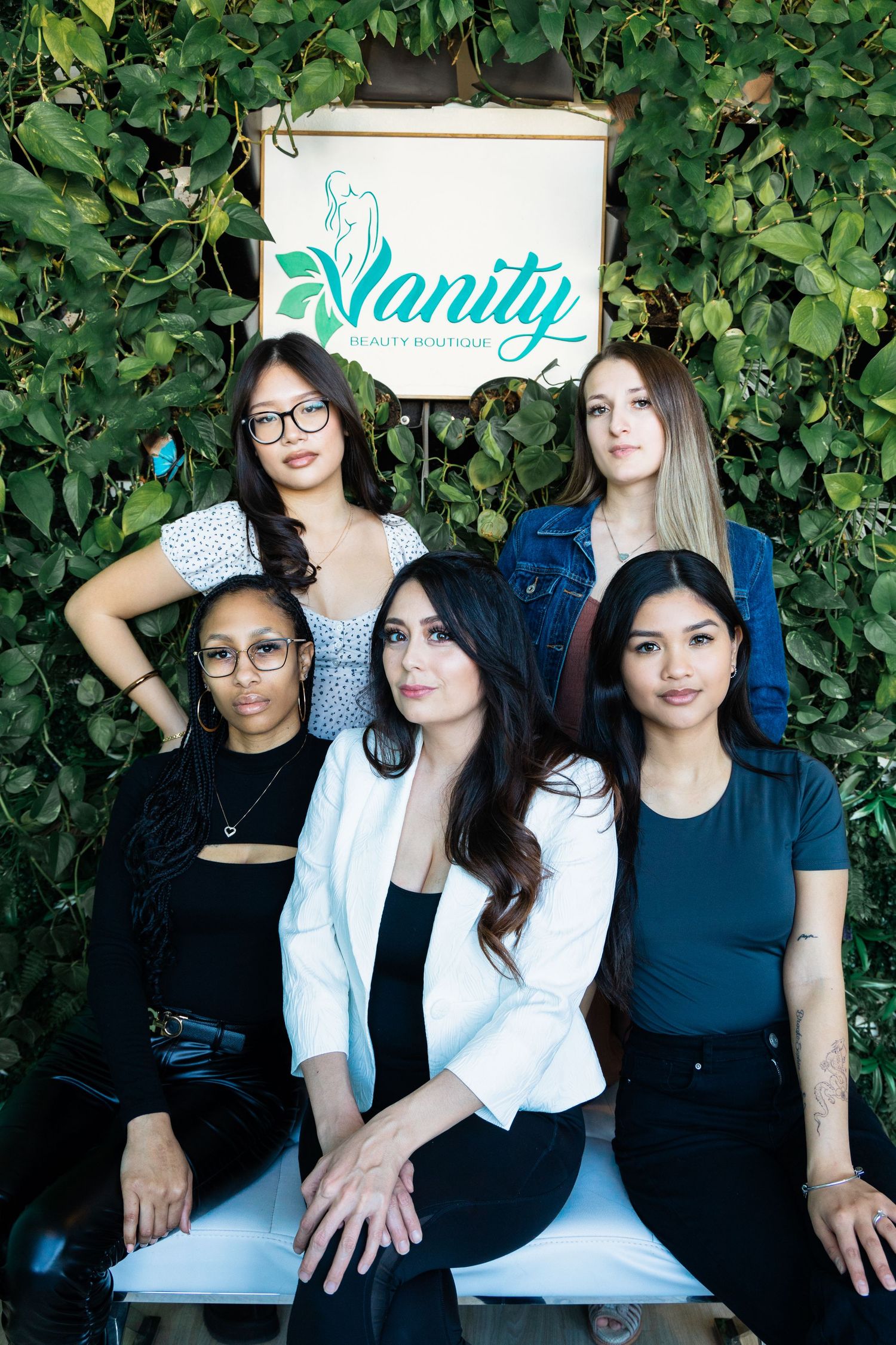 Five women pose together in front of a green ivy wall with a Vanity Beauty Boutique sign.