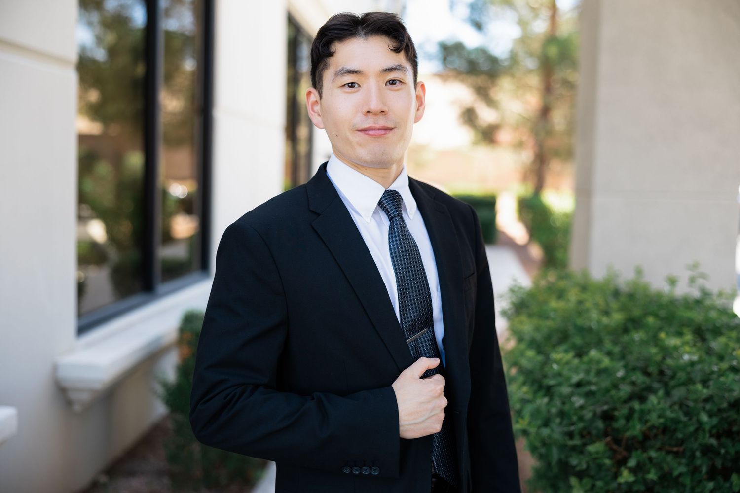 Young Asian man in black suit and tie poses confidently outdoors near building with greenery.